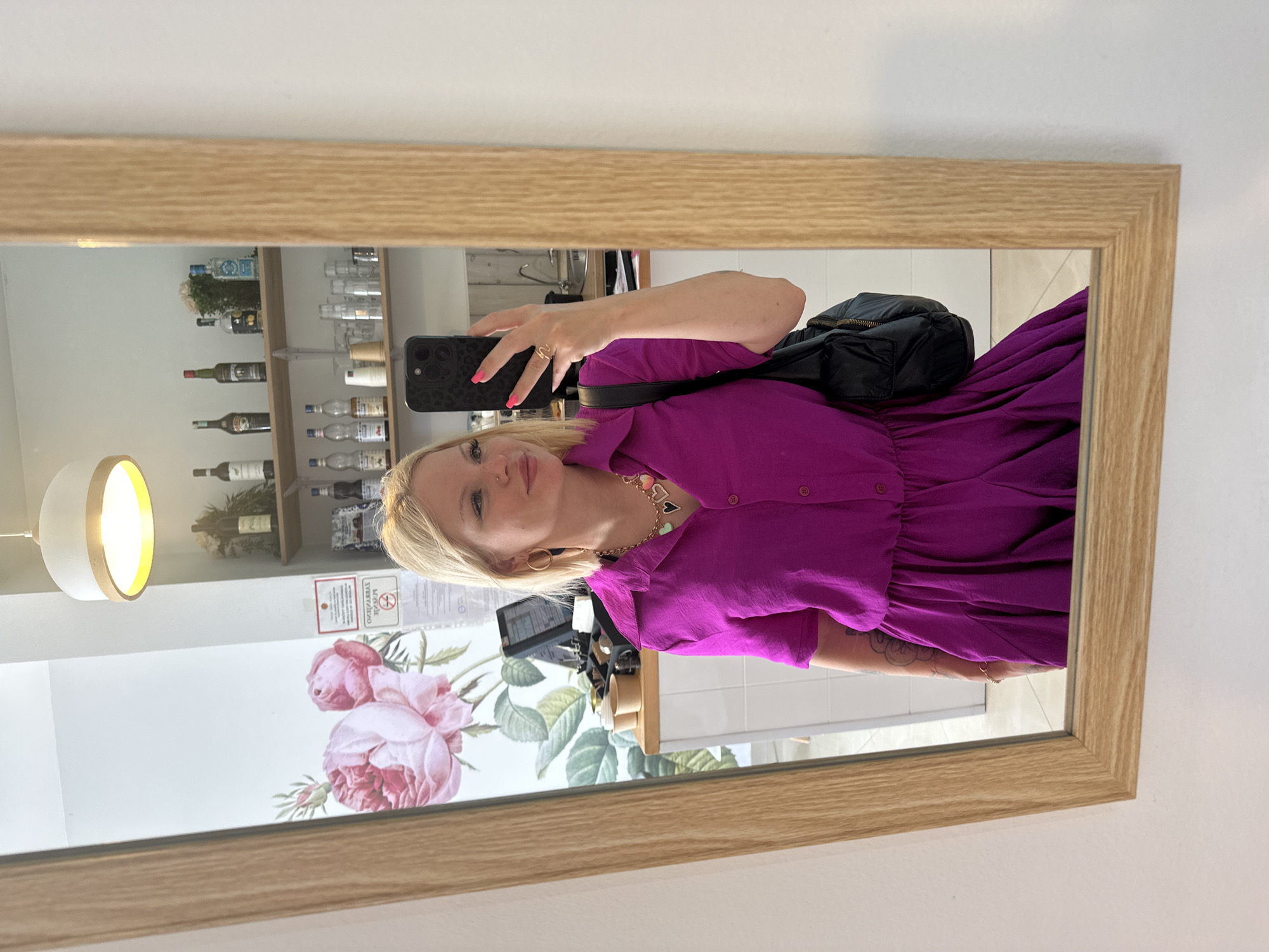 The image captures a blonde woman in Budva, Montenegro, taking a mirror selfie inside what appears to be a commercial establishment, possibly a cafe or bar. The woman, positioned centrally, is reflected in a rectangular mirror with a light wooden frame. She wears a magenta, short-sleeved, collared dress with a cinched waist. Her left hand, adorned with bright pink nail polish and a gold-colored, wave-like ring, holds a black smartphone with a textured black case, obscuring part of her face. She has gold hoop earrings, a small nose piercing, and a necklace featuring multiple heart-shaped charms in various colors including black, light green, pink, and gold. A tattoo with a circular design is partially visible on her left forearm. A black quilted shoulder bag or small backpack is visible on her right side.
The reflected background reveals the interior of the establishment. To the woman's left, a wall features a large, detailed decal or painting of pink roses and green leaves. Below this, a light-colored counter holds two light brown disposable cups and a black point-of-sale system. A white sign with red text and a "no smoking" symbol is mounted on the wall to the right of the floral decor, showing partially visible text that reads "ЗАБРАЊЕНО ПУШЕЊЕ" (No Smoking). To the woman's right, two light wooden shelves are mounted on a white wall. The upper shelf displays several bottles, including dark green wine bottles and various liquor bottles. The lower shelf holds a selection of clear glass cups and several smaller bottles, possibly containing syrups. Above, a modern ceiling light fixture with a white conical shade and a light wooden band emits a warm yellow light. In the far background, reflected behind the woman's head, a metal sink and faucet are visible, suggesting a utility or preparation area. The flooring appears to be light-colored tiles.