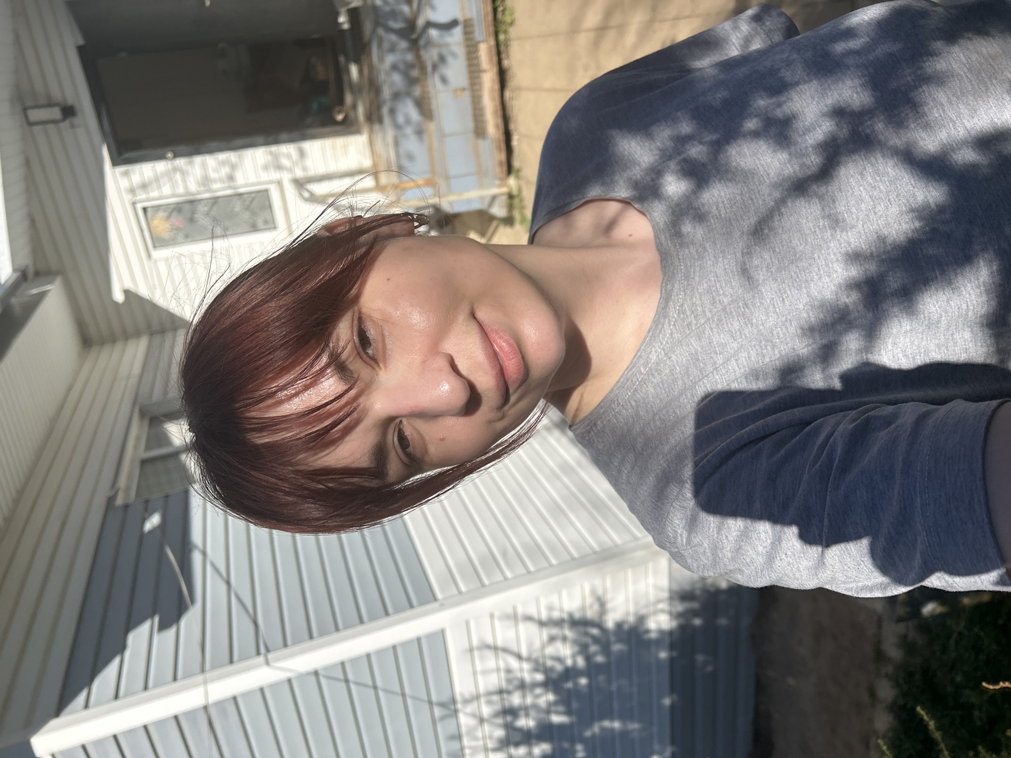 A woman with auburn hair is photographed outdoors on a sunny day. She is wearing a gray t-shirt and facing the camera. Behind her is a light blue and white wooden house with visible windows. The lighting is bright, with shadows from nearby trees falling on the woman and her surroundings. The location cannot be determined as Фоки, Russia with the available information.