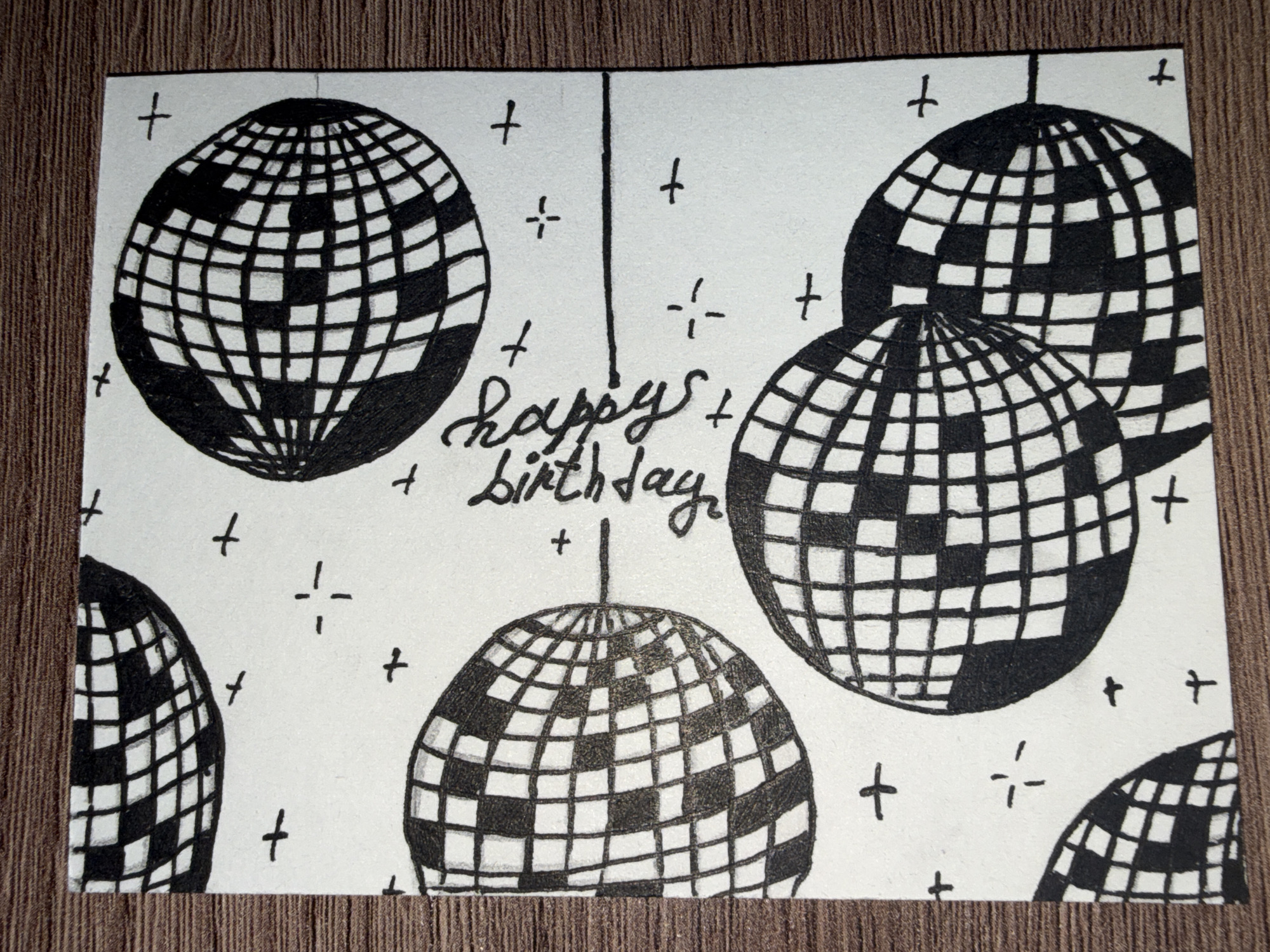 The image is a hand-drawn illustration on a white card with a textured wooden background. The illustration depicts several disco balls scattered across the card, with small plus signs and star-like shapes also present. In the center of the card, the words "happy birthday" are written in cursive. The disco balls are rendered in black and white, with a grid pattern representing their mirrored surfaces. There are no people or any indications of a specific location in Rayevskiy, Russia.