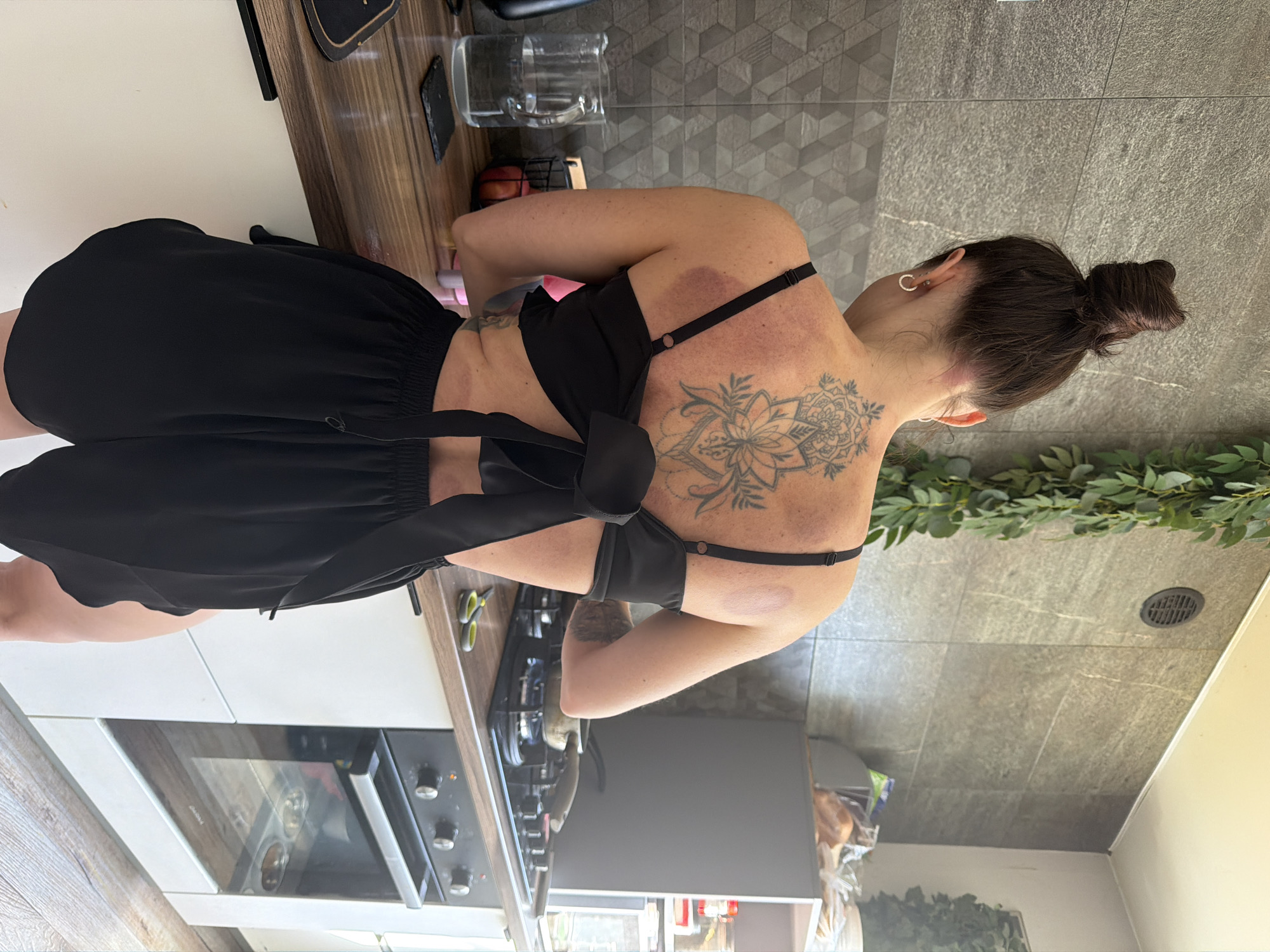A woman with dark hair in a bun stands in a kitchen. She is wearing a black top and shorts, and has a large floral tattoo on her upper back. There are several circular bruises visible on her back and upper arms. The kitchen has light-colored countertops and grey tile backsplash. A stove with pots and pans is visible, as well as kitchen appliances. There is greenery draped along a wall in the background. The location cannot be determined as Perm, Russia from provided context.