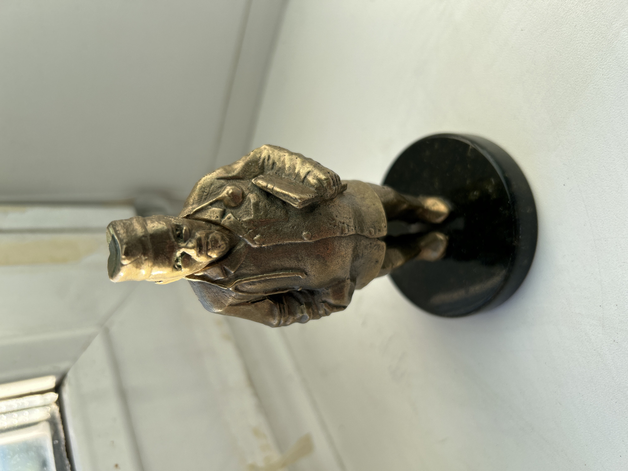 A small bronze figurine of a doctor stands on a black circular base on a white, textured surface. The figurine depicts a male figure wearing a cap, a coat, and a stethoscope around his neck. He holds a book in his left hand and has his right hand in his coat pocket. The figurine is positioned in front of a window with white mullions, partially obscured by light and glare. The background is a plain, light-colored wall. The overall scene is brightly lit, suggesting it is daytime. There is no identifiable context or specific location within the city of Tomsk, Russia, provided by the visual information alone.