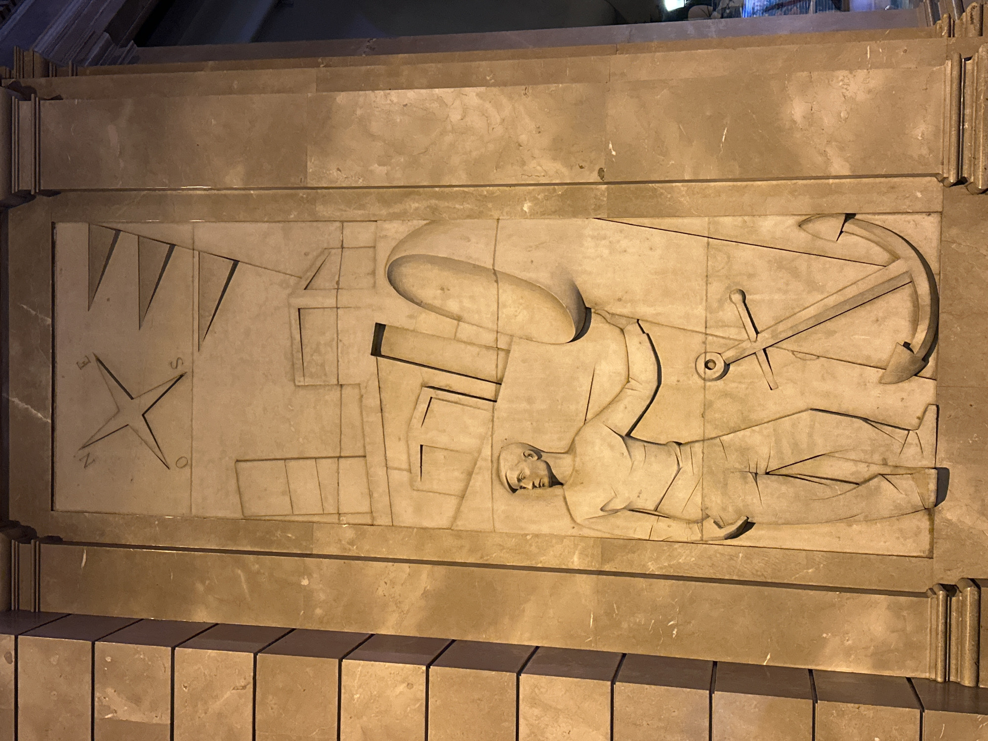 The image depicts a stone bas-relief panel. The scene carved in the panel includes a compass rose, a maritime sextant, a man lying prone in uniform, and a naval anchor. The panel is a light beige color with some variations in tone and veining. The panel is framed with stone borders and is located above stone block steps. This is a detail of the monument to Cristopher Columbus (Colom) in Barcelona, Spain.
