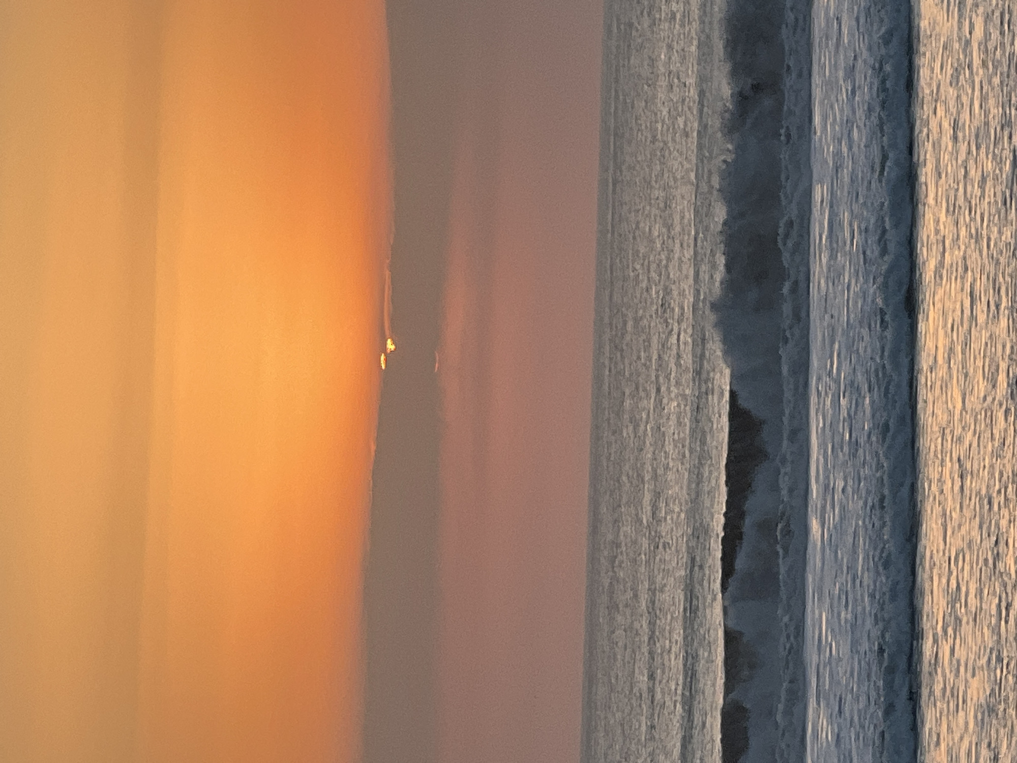 The image depicts a serene ocean sunset in Kuta, Indonesia. The sky is ablaze with vibrant orange and pink hues, with the sun partially obscured by a thin layer of clouds near the horizon. The ocean below is calm, with gentle waves rolling towards the shore. The water reflects the warm colors of the sunset, creating a tranquil and picturesque scene. The image captures the natural beauty of Kuta's coastline during sunset.