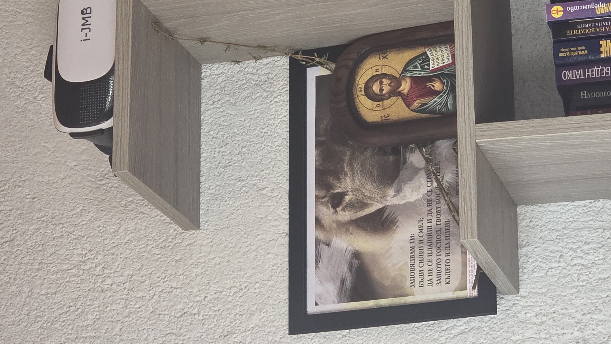 This image captures a close-up of a wall-mounted shelving unit, likely in a domestic setting in Bania, Bulgaria. The shelves are made of a light wood-colored material and hold various personal items.
On the underside of the top-left shelf, a white and black device labeled "i-JMB" is mounted, possibly a small speaker or security camera.
Beneath the right-hand shelf, a vertically oriented framed picture is displayed. The picture features the head of a large cat, possibly a lion, in the lower section, while the upper section contains Cyrillic text. This text, likely Bulgarian, reads: "ЗАПОВЯДВАМ ТИ: / БЪДИ СИЛЕН И СМЕЛ; / ДА НЕ СЕ ПЛАШИШ И ДА НЕ СЕ СТРАХУВАШ; / ЗАЩОТО ГОСПОД, ТВОЯТ БОГ Е С ТЕБ / КЪДЕТО И ДА ИДЕШ." (I COMMAND YOU: BE STRONG AND COURAGEOUS; DO NOT BE AFRAID AND DO NOT BE DISMAYED; FOR THE LORD YOUR GOD IS WITH YOU WHEREVER YOU GO.)
Above this framed picture, a small Orthodox Christian icon of Jesus Christ is visible, set in a wooden frame. The icon clearly depicts Jesus with a halo, and the Greek abbreviations "IC" and "XC" (for Jesus Christ) are visible near his head, along with "O ΩΝ" (The Existing One) in the halo. A small, dried plant sprig is draped between the icon and the shelf above.
On the shelf to the right, a stack of books is visible. Several titles are in Cyrillic, presumably Bulgarian: "Мпредимство" (Advantage), "ТА НА БОГАТИТЕ" (To the Rich), "ИЛА НА ПАРИТЕ" (Power of Money), and prominently, "БЕДЕН ТАТКО" (Poor Father), which is part of the popular "Rich Dad Poor Dad" series. Another book has "НАПОЛЕОН" (Napoleon) on its spine and "www.anhira.com" and "здраве, успех, свобода" (health, success, freedom) visible on its cover.
The background wall has a textured, off-white finish. No specific activity is taking place; the image is a static capture of the display.