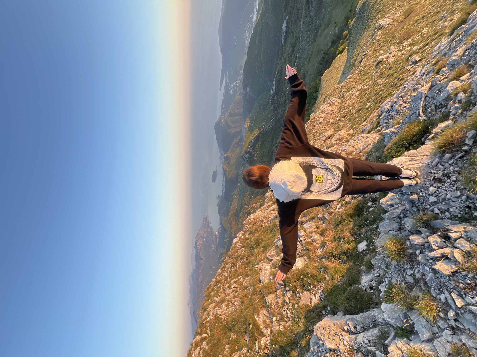 A woman stands at the edge of a rocky cliff in what appears to be a mountainous region. She wears a black hooded sweatshirt with a white patch on the back, dark-colored pants, and light-colored shoes. Her arms are outstretched, mimicking a bird in flight. She wears a white hat. In the background, there is a vista of mountains, water, and a pale sky. The location is near Dolovi, Montenegro. The landscape is rocky and partially covered with grasses.