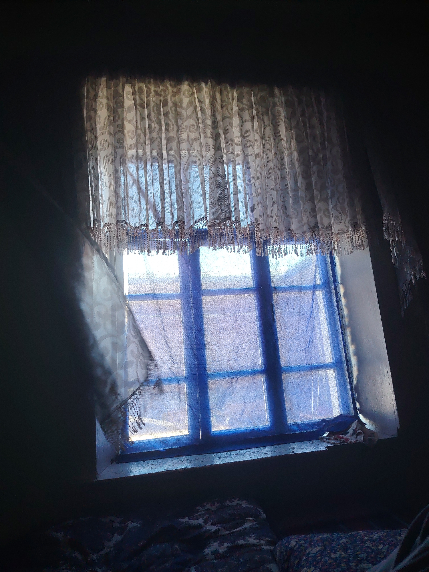 The image shows a window with blue-tinted glass panes and white lace curtains. The window is framed by a dark interior, suggesting it is inside a building. Sunlight filters through the curtains, casting shadows and illuminating parts of the window and the wall. On the windowsill, there are a few crumpled pieces of fabric or paper. Below the windowsill, a patterned surface, likely bedding, is visible. The overall impression is of a dimly lit room with a window as the primary source of light. The location in Almachi, Uzbekistan, is not explicitly depicted in the image itself, but this context provides a geographical setting for the scene.