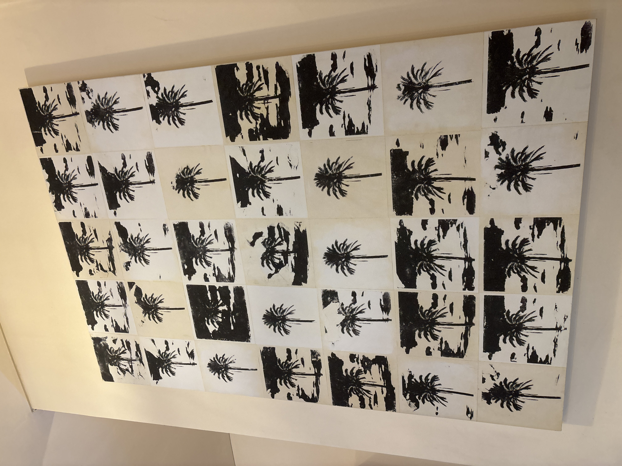 A large, multi-panel artwork featuring stylized palm tree prints is prominently displayed on a light-colored wall in an interior setting. The artwork is composed of numerous square or rectangular tiles, arranged in a grid approximately six rows by seven columns, though the extreme edges are slightly out of frame. Each tile showcases a graphic print of a palm tree, depicted in black or dark brown against an off-white or cream background, or as light silhouettes against a dark, textured background.
The palm trees are generally oriented horizontally, with their fronds extending to the left and a trunk to the right. The prints exhibit a raw, expressive quality, suggestive of linocut or woodcut techniques, with visible brushstrokes, textures, and distressed edges that add to their artistic character. This alternating pattern of light-on-dark and dark-on-light creates visual variation and rhythm across the entire piece.
The artwork is mounted slightly off the cream-colored wall, creating a subtle shadow that gives it depth. The lighting in the room is even and soft, indicating an indoor environment. There is no activity taking place, as the image captures a static art installation. The recurring palm tree motif is highly relevant to the provided location context of Kuta Selatan, Indonesia, a region known for its tropical landscapes. There is no visible text on the artwork or the wall.
