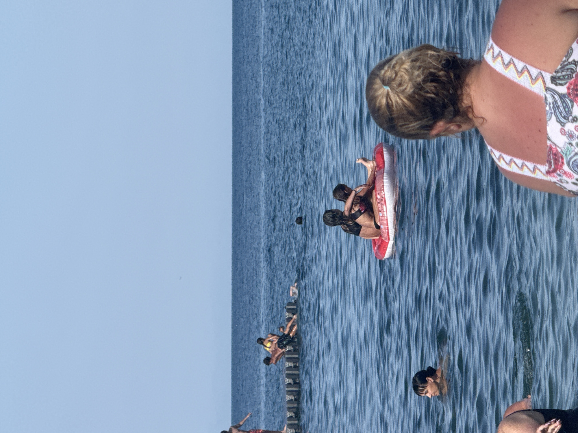 This is a vertically oriented, slightly blurry photograph taken on a sunny day at the beach. The scene is dominated by the deep blue of the sea, with a clear, light blue sky above. Several people are visible in the water. In the foreground, a woman's head and shoulders are visible, seen from behind, wearing a patterned swimsuit. She appears to be looking out at the sea. In the middle ground, a group of people are on an inflatable raft. One person is lying down, and another two are sitting. Further back, a group of people are on a wooden pier or jetty. Some are sitting or lying on it, while others are in the water near it. There is a sense of leisure and recreation. The water is calm with gentle ripples. The time of day appears to be midday or early afternoon given the bright light. The location is a beach in Partenit, Ukraine. There is no visible text in the image.