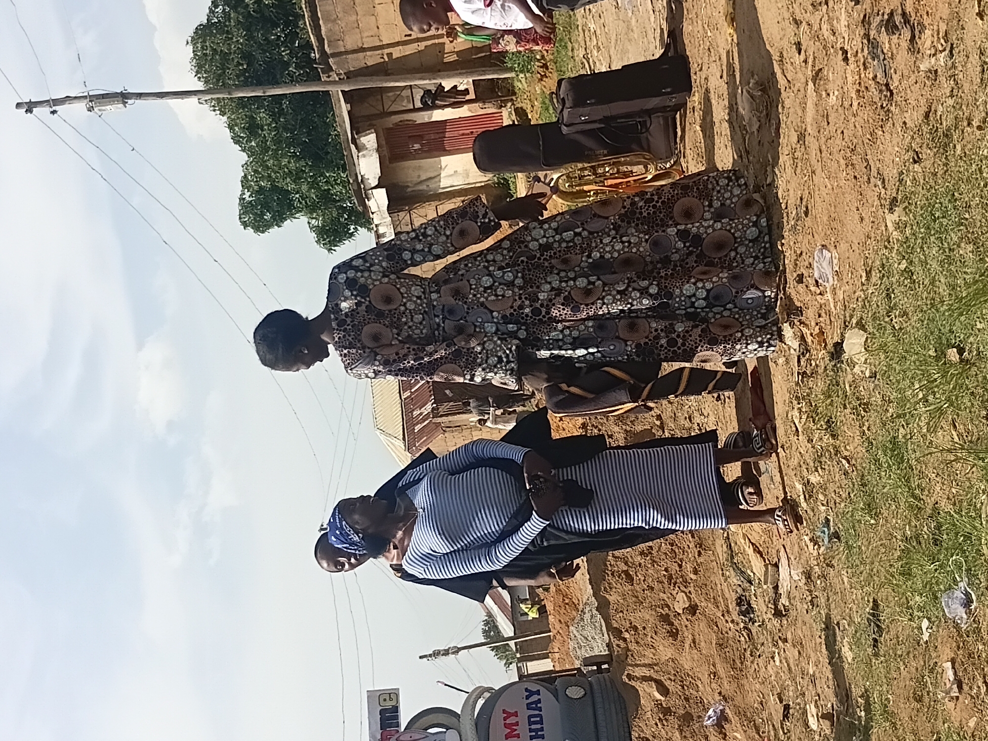 In Jos, Nigeria, a group of people are gathered on a dusty, unpaved street near a building. A woman in a long, patterned dress stands with her back to the camera, facing two other individuals. To her left, a woman wearing a blue headscarf and a striped dress appears to be speaking. Behind her, a person in a black garment is partially visible. In the foreground, a brass musical instrument case and a suitcase are placed on the ground. A young boy in a white shirt stands to the right, looking towards the musical instruments. In the background, a sign with the word "WELCOME" and "MY BIRTHDAY" is visible, along with corrugated metal roofs and brick walls. The ground is littered with debris, and the sky is partially cloudy.