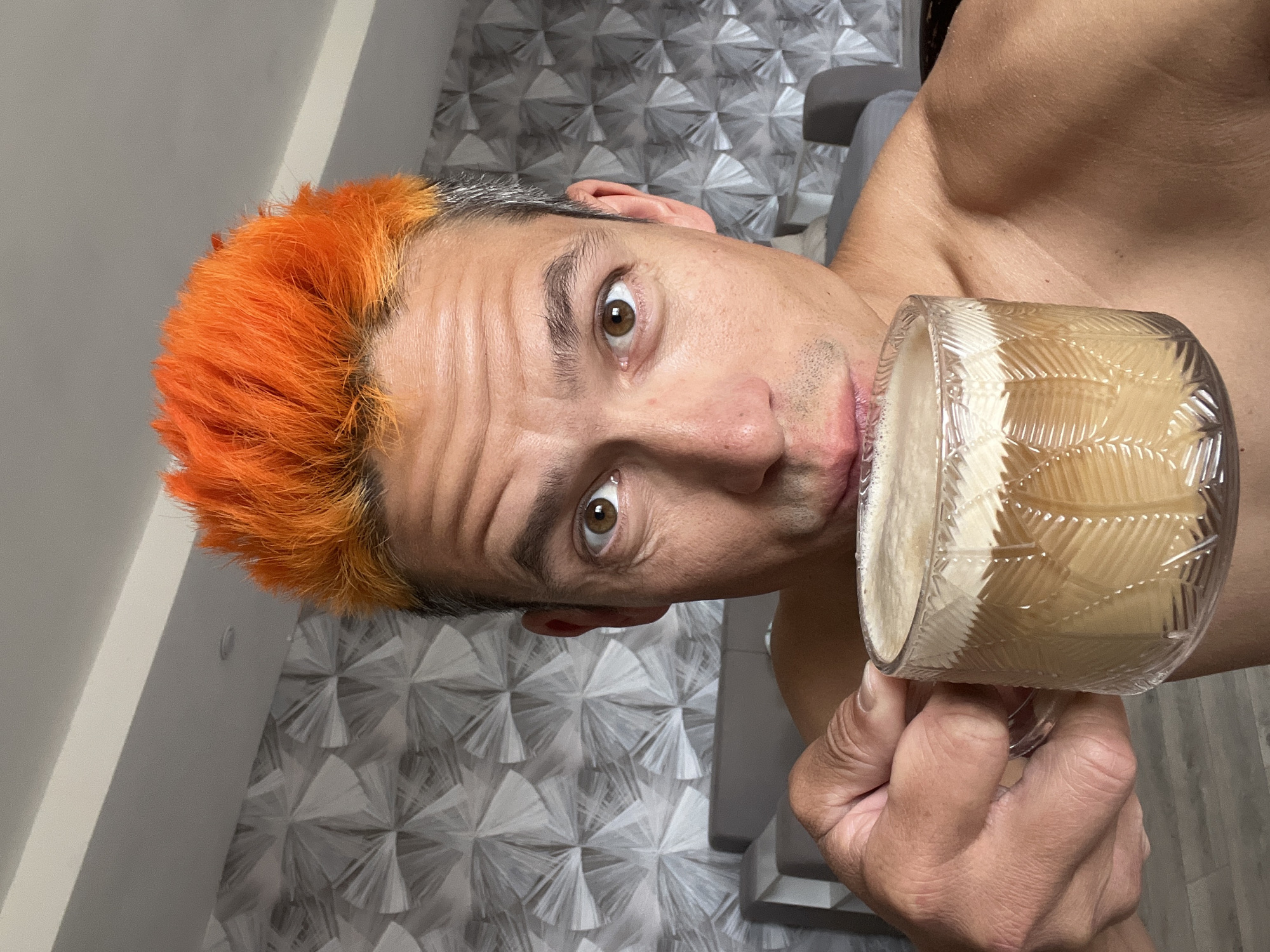 This image, taken in Chaikovsky, Russia, features a close-up of an adult male. He has vivid orange, spiky hair, with visible grey growth at the roots on the sides of his head. His brown eyes are open wide and directed towards the camera, with a slight furrow to his brow. His bare shoulders and upper chest are visible, showing a light tan line on the right shoulder. The male is holding a clear glass mug with a textured, patterned surface in his right hand. The mug contains a light brown liquid topped with a thick layer of white foam, suggesting a beverage such as coffee with cream or froth. He is positioned as if taking a sip from the mug, with his lips near the rim. The background consists of a wall covered in a distinctive three-dimensional wallpaper pattern composed of numerous intersecting light grey and white geometric shapes. Blurred grey furniture is partially visible in the lower background, and a white ceiling with a recessed light fixture is visible in the upper left.