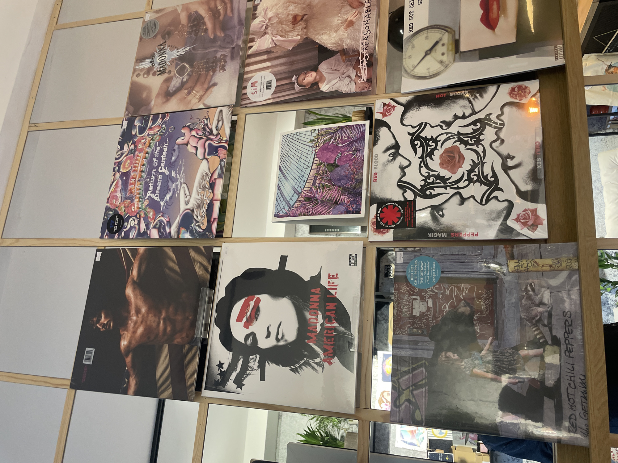 The image shows a display of vinyl records in a rack. The albums visible include Madonna's "Madonna", Sia's "Reasonable Woman", a clock and a pair of red lips, Red Hot Chili Peppers' "Blood Sugar Sex Magik", "Return of the Dream Canteen", "The Getaway", Madonna's "American Life", and an album named "Red Blood". The location is not discernible from the image, nor is the city of Novi Sad, Serbia. The rack itself is made of light-colored wood.