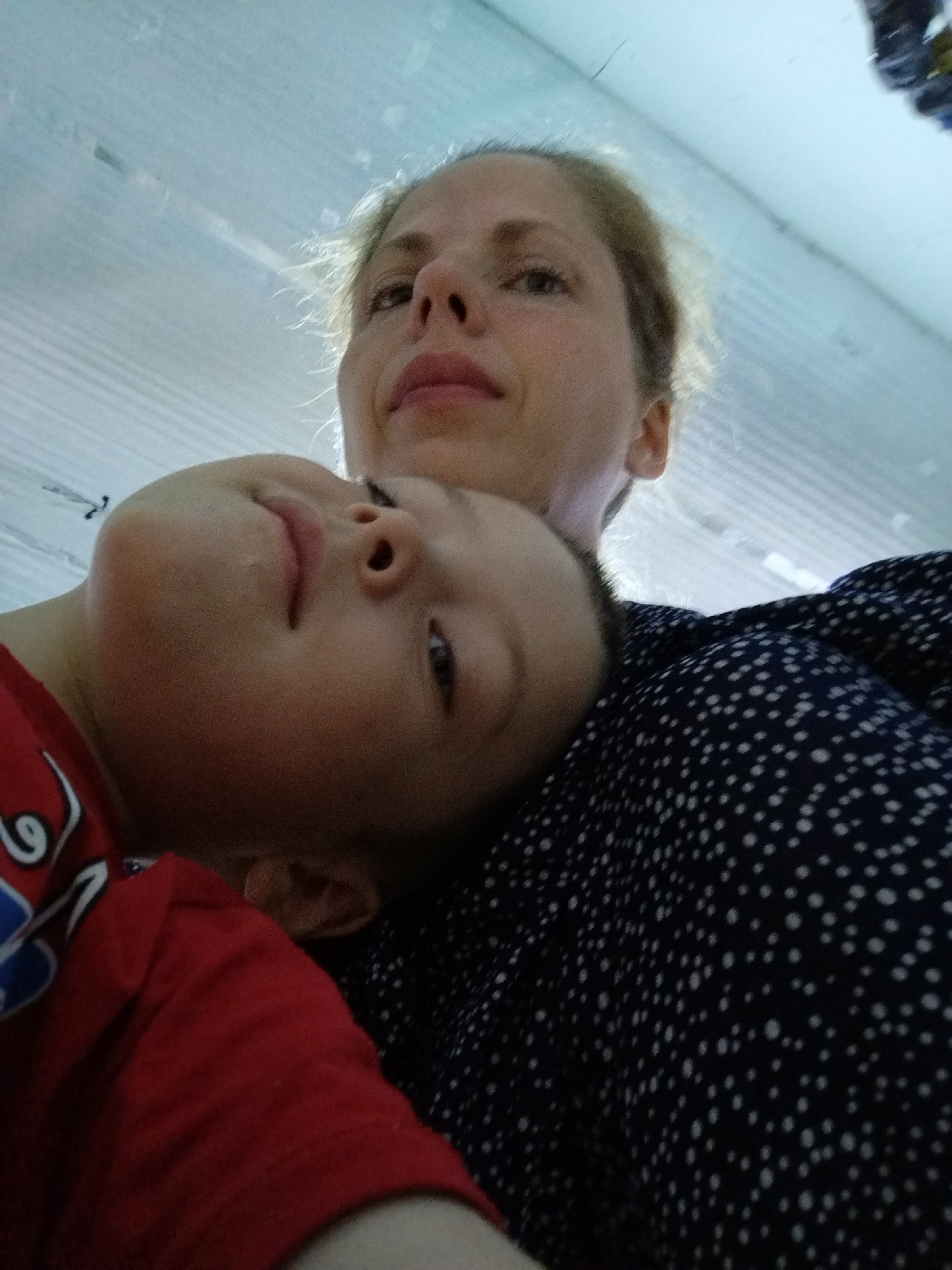 A low-angle, close-up, outdoor selfie shows a blonde woman's face in the upper right and a young child's face in the lower left. The woman is looking up and slightly away from the camera, with her mouth open. Her hair is light blonde and windswept. The child is resting their head on the woman's shoulder or chest, with their eyes closed and mouth slightly open. The child is wearing a red t-shirt with some blue detailing. The woman is wearing a dark blue shirt with a white polka-dot pattern. The background appears to be a light blue, textured surface, possibly a ceiling or a wall. The lighting is bright and diffused.