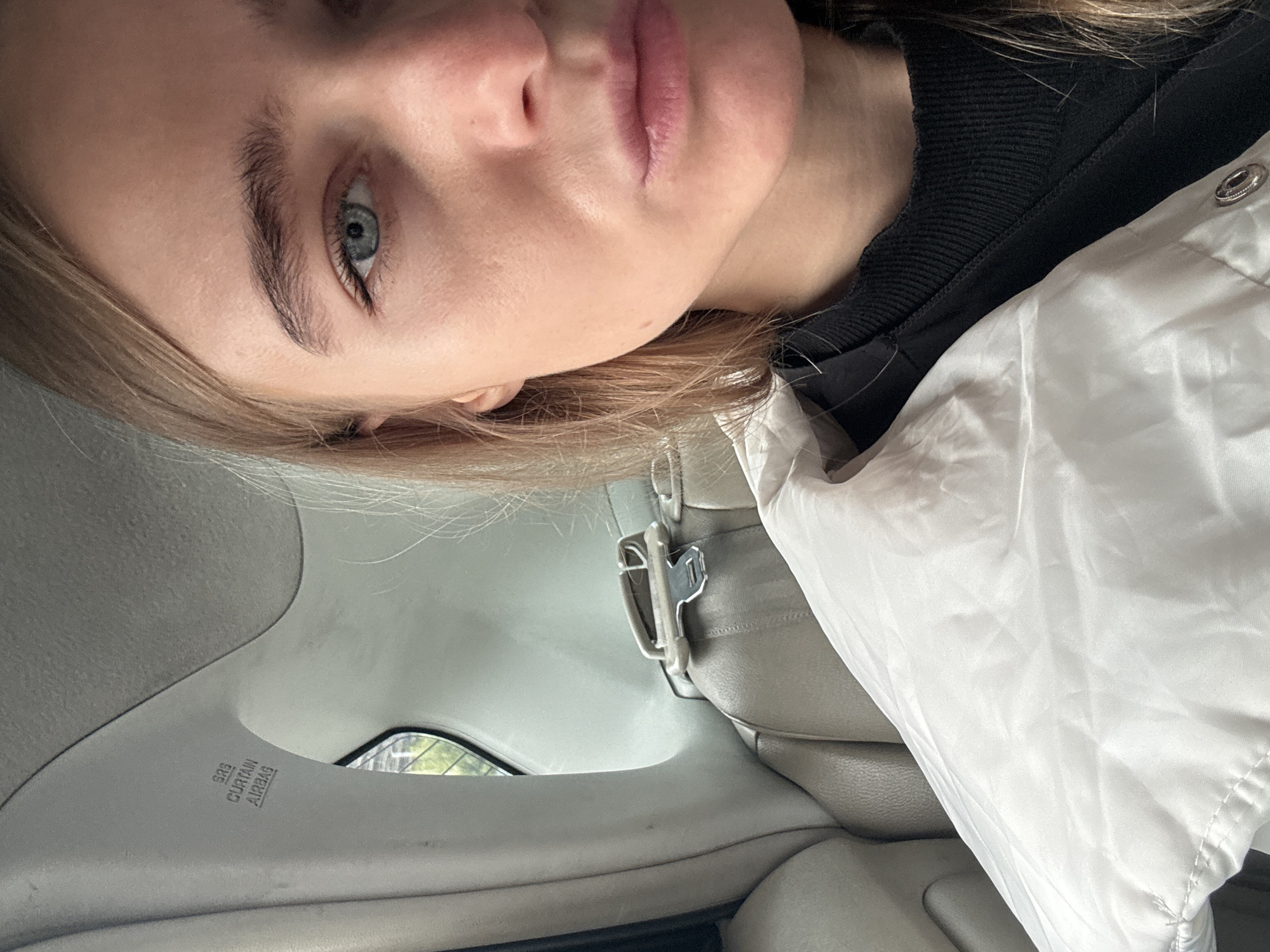 A close-up, rotated image shows a person with fair skin, blonde hair, and light blue eyes, positioned in the passenger seat of a car. The person is wearing a black top and a white jacket. The car interior is visible, including a gray leather seat, seatbelt hardware, and a portion of the car's frame with the inscription "SRS CURTAIN AIRBAG". Through the car window, a glimpse of foliage indicates an outdoor environment. The location and specific details of the city remain unknown.