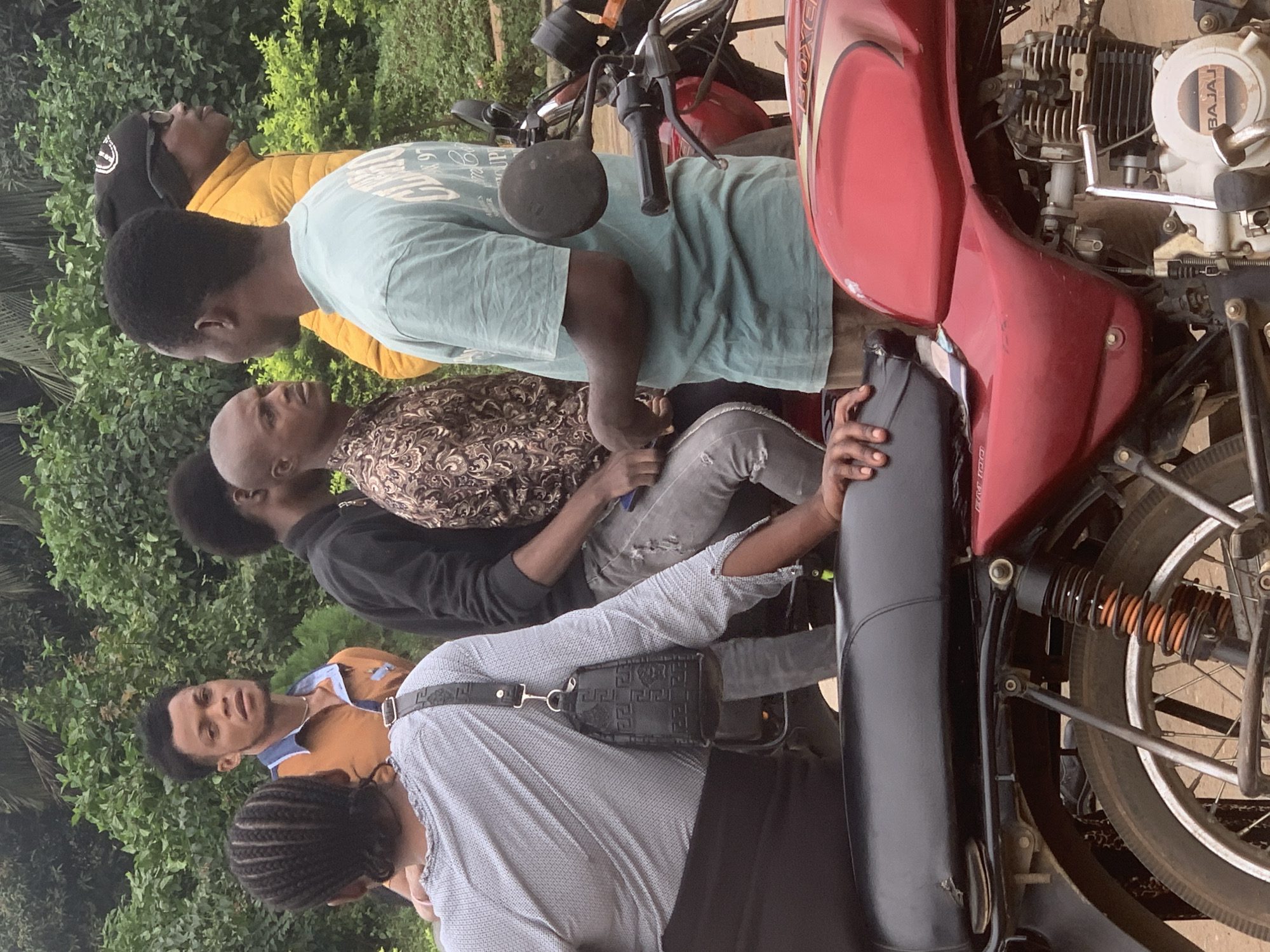 The image depicts a group of individuals gathered around a red motorcycle in Shagamu, Nigeria. In the foreground, a person is seated on the motorcycle, facing away from the camera, wearing a grey shirt and black pants. A black bag with a patterned design is attached to their shoulder. To the right of the motorcycle, another person wearing a light blue t-shirt and brown shorts stands, conversing with a person in a patterned shirt and grey jeans who is seated on the motorcycle. Behind them, a man in a yellow jacket and a black beanie, along with another man in a brown shirt, are also present. The background consists of dense green foliage, suggesting an outdoor setting. The overall scene appears to be a casual gathering or waiting area.