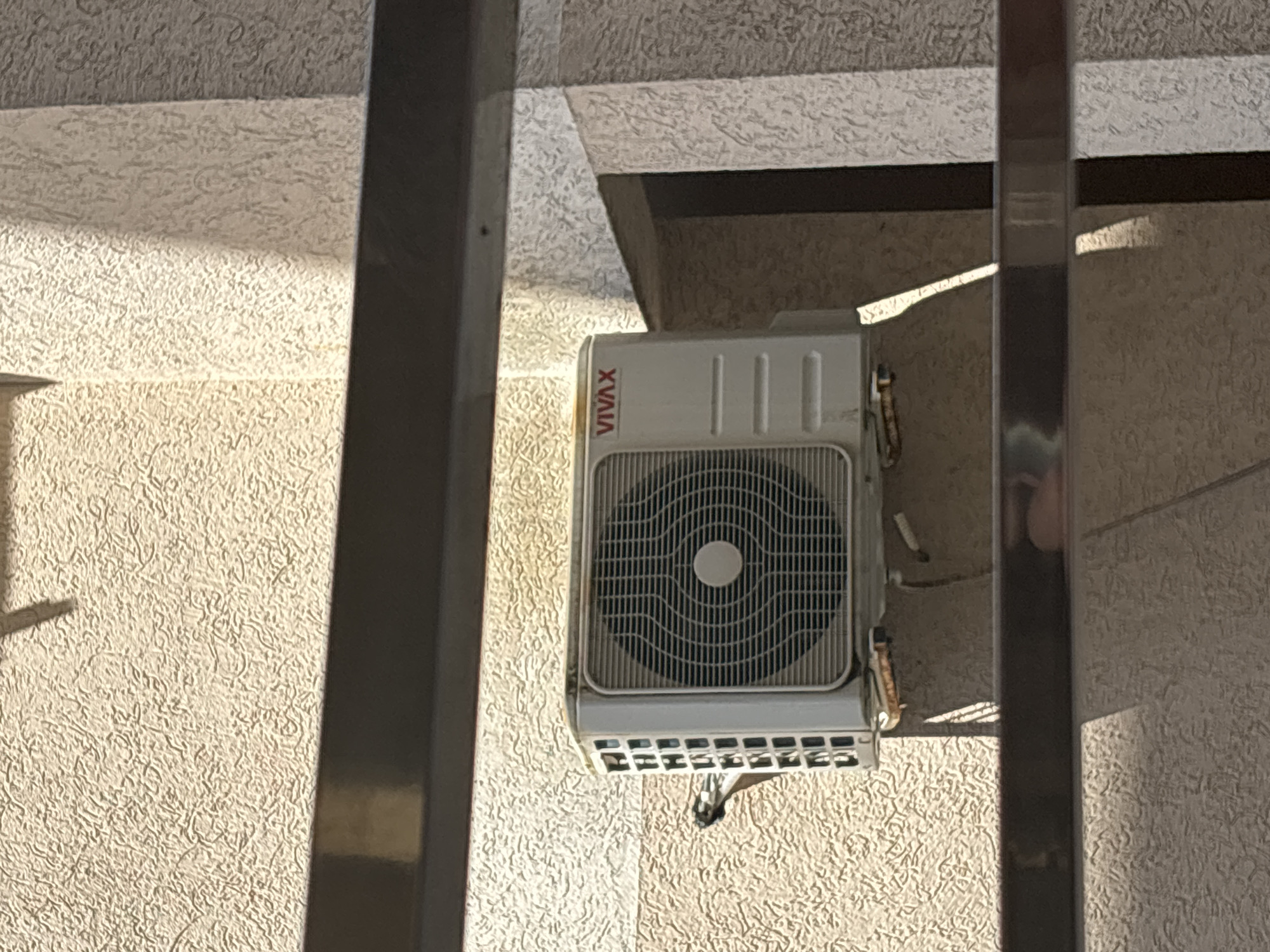 A white VIVAX air conditioning unit is affixed to a textured beige exterior wall. The unit is situated between two dark, vertical metal bars that appear to be part of a railing or structure. The location is Budva, Montenegro.