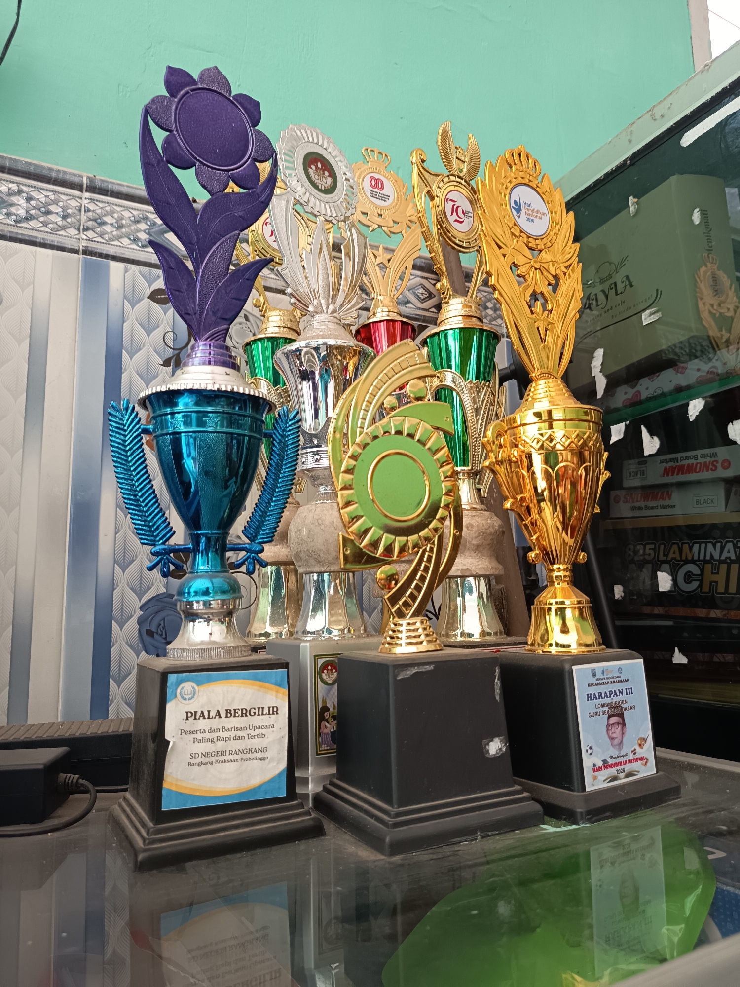 In the city of Widoro, Indonesia, a collection of trophies sits on a reflective surface, possibly a table, with a green wall in the background. The trophies are of various sizes and designs, with some featuring golden metallic finishes and others a striking blue. One trophy, adorned with a purple floral element, stands out at the front left. In front of the trophies are labels with text in Indonesian, one of which reads "PIALA BERGILIR Peserta dan Barisan Upacara Paling Rapi dan Tertib SD NEGERI RANDANGGIRANG KRAKSAAN-PROBOLINGGO". Another label, also in Indonesian, states "HARAPAN III LOMBA KREASI GURU SEKOLAH DASAR Hari Pendidikan National 2023" and includes a portrait of a man wearing glasses. The trophies appear to be arranged in a display, likely in an office or educational institution. The scene is well-lit, suggesting it was captured during the daytime.