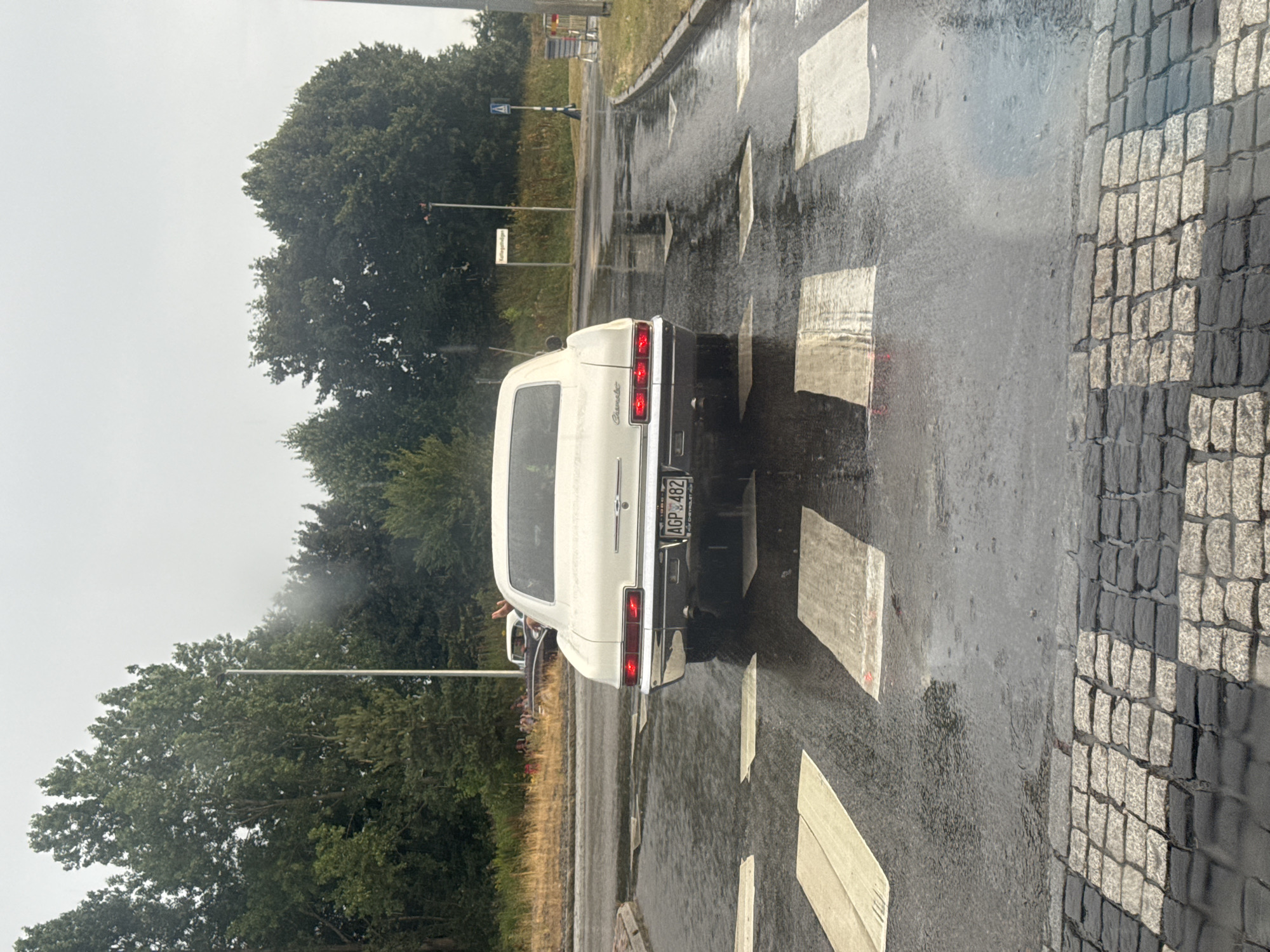 The image depicts a white Chevrolet Impala from the mid-1960s, with a Swedish license plate reading "AGP 482", parked on a wet road at a pedestrian crossing. The car is facing away from the camera, with its taillights illuminated. It is raining, as evidenced by the wet road surface, reflections, and visible raindrops. In the background, there are trees, streetlights, and a sign that reads "Karlsgatan". The road is paved with cobblestones in the foreground and asphalt further back. The sky is overcast, suggesting a cloudy or rainy day. There are no people clearly visible interacting with the car, though a person's hand is raised in another car in the distance.