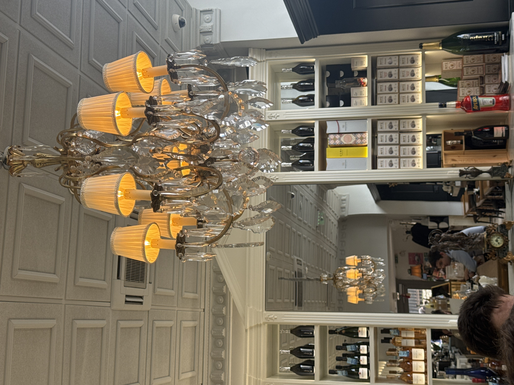 Here is a forensic description of the image:
**General Impression:** The image captures an indoor scene with a mix of architectural elements and human presence, likely within a high-end commercial establishment. The composition focuses on a decorative chandelier and a mirrored wall reflecting the interior space.
**Specific Details:**
* **Foreground:** A prominent, ornate chandelier with gold-colored metal framework and multiple lamps with pleated fabric shades is suspended from the ceiling. Transparent crystals adorn the chandelier's structure.
* **Background:** Behind the chandelier, a wall with recessed rectangular panels is visible. To the right, a mirrored wall reflects the interior of what appears to be a bar or restaurant.
* **Mirror Reflection:** The mirror reflects an extended view of the interior, including another matching chandelier further down the hall. A wine rack with visible bottles and boxes of "Ritz" crackers is adjacent to the mirror.
* **People:** A person with brown hair is slightly visible on the right of the mirrored section. A second person can be seen in the reflection working behind a counter.
* **Merchandise:** In the mirrored reflection, several bottles and containers are visible, including recognizable "Aperol" and "Ritz" branded products.
* **Environment:** The setting appears to be an upscale establishment, possibly a restaurant or hotel lounge. The lighting is warm due to the chandelier and ambient light.
* **Additional Details:** A security camera can be seen near the ceiling. There is a vintage air conditioning unit on the panelled wall.
**Location:**
It is difficult to ascertain with certainty that the picture was taken in Sofia, Bulgaria. Further information is required to confirm that the scene is within Sofia.