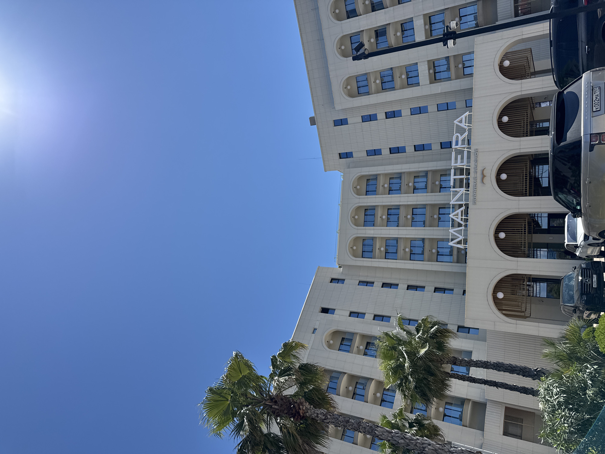 The media file shows a contemporary hotel building with a bright blue sky and lush palm trees in the foreground. The hotel, identified by the text "MANTERA SUPREME SEASIDE" visible on its facade, has a modern architectural design with multiple levels, balconies, and numerous windows. The sun is shining brightly, casting a glare on the left side of the image, indicating it is daytime. The presence of palm trees suggests a warm climate, and the clean, well-maintained appearance of the hotel implies it is a resort or a high-end establishment. Parked cars are visible next to the building, including a silver Range Rover with a license plate that reads "K002MA 197". The overall impression is one of a sunny, pleasant day at a seaside location. The image is oriented vertically.