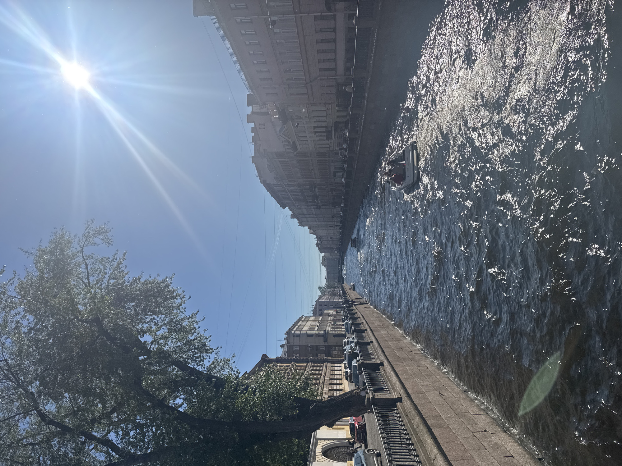 The image captures a sunny day in Saint Petersburg, Russia, along a canal. The canal's banks are lined with stone walls and ornate iron railings. On the left bank, trees overhang the canal, and a row of buildings and parked cars are visible. The right bank features a long stretch of multi-story buildings with architectural details typical of the city. Several small boats are navigating the canal, with one boat carrying passengers prominently in the foreground, creating ripples on the water's surface. The sky is clear blue, with the sun shining brightly, casting a glare and creating sparkling reflections on the water. The overall impression is one of urban beauty and activity on a pleasant day.