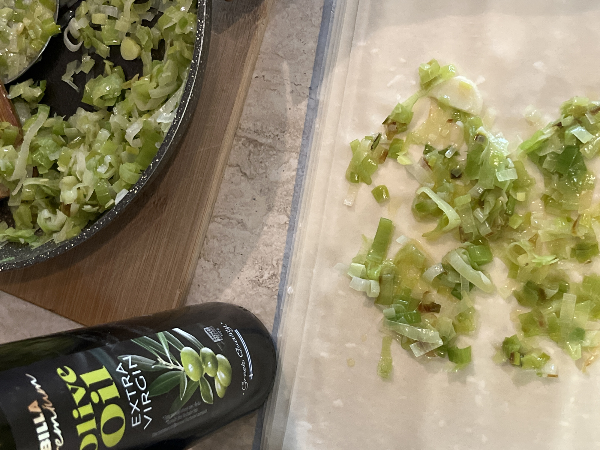 The image depicts a culinary scene indoors. A frying pan filled with sautéed leeks sits on a wooden cutting board. A bottle of "Billa Premium Olive Oil Extra Virgin Greek Quality" lies near the bottom left of the frame. Some sautéed leeks are spread across a light-colored, rectangular surface. The location and Sofia, Bulgaria are not evident in the image.