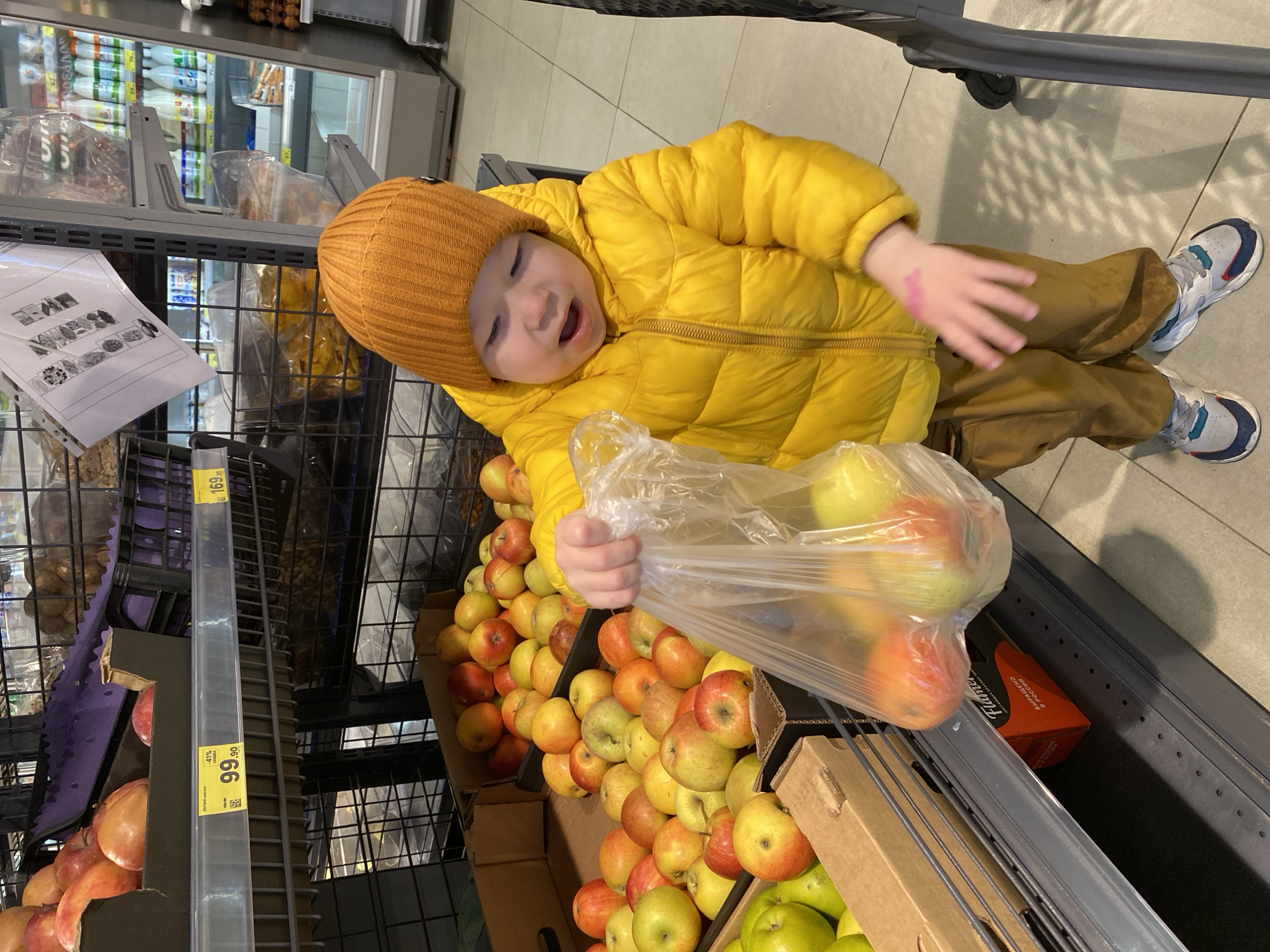 A child wearing a yellow puffer jacket and a mustard-colored knit hat sits in a shopping cart in what appears to be a supermarket. The child holds a plastic bag filled with apples. The apples are displayed in open cardboard boxes within the shopping cart. Price tags are visible on nearby shelves, indicating the prices of other items in the supermarket. The packaging of "Fitomed" which is produced in Russia can be seen beside the apples. The location is within a supermarket in Sochi, Russia.