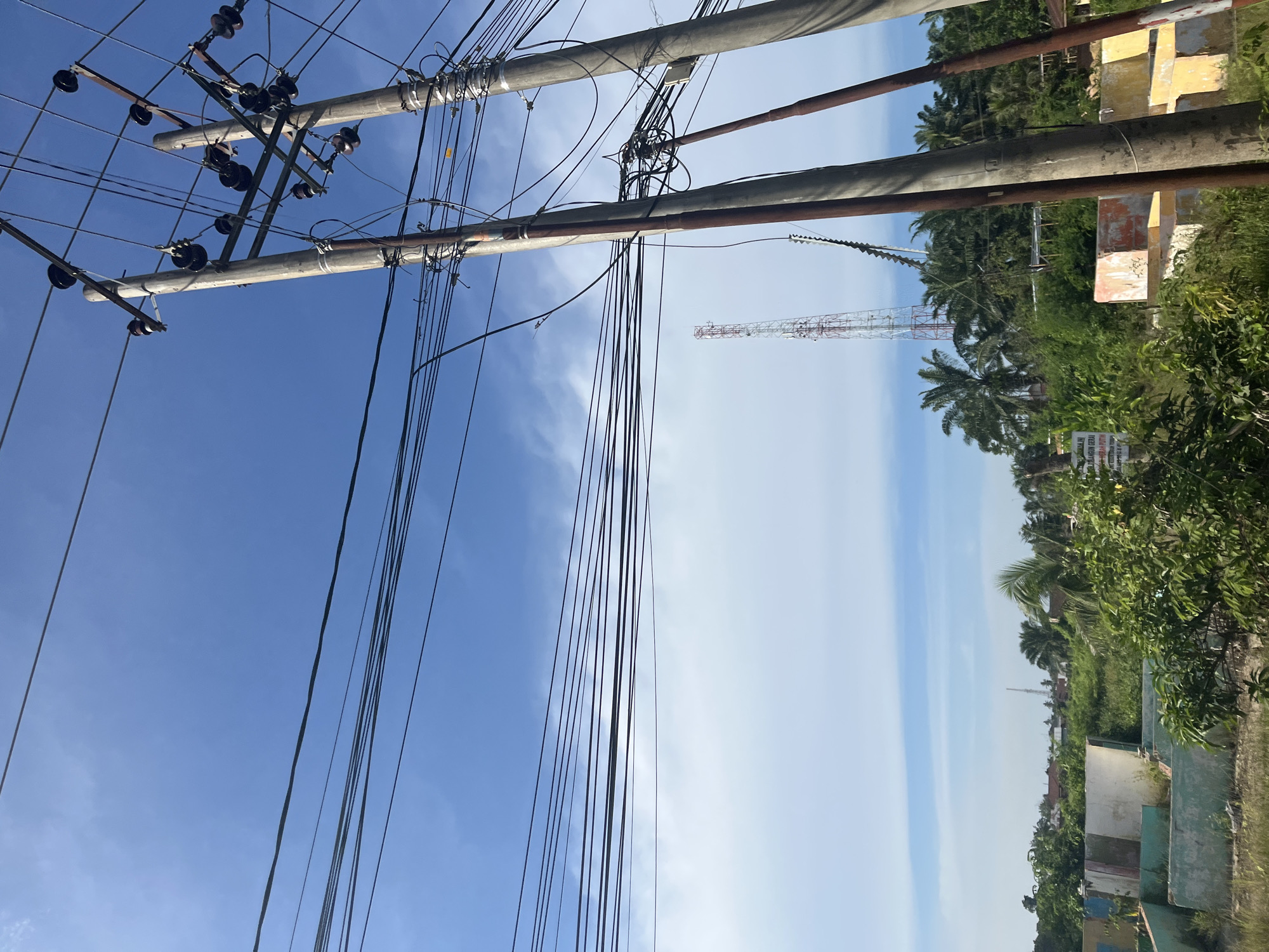 This is a ground-level, outdoor, daytime photograph taken in Tanahterban, Indonesia. The image captures a view of utility poles with numerous overhead wires against a clear blue sky with some wispy white clouds. In the background, a tall, red and white communication tower is visible, surrounded by lush green vegetation, including palm trees and smaller trees. Some partially constructed or dilapidated concrete structures are also present in the mid-ground, with signs of paint in various colors. The overall scene depicts a rural or semi-urban environment with infrastructure and natural elements.