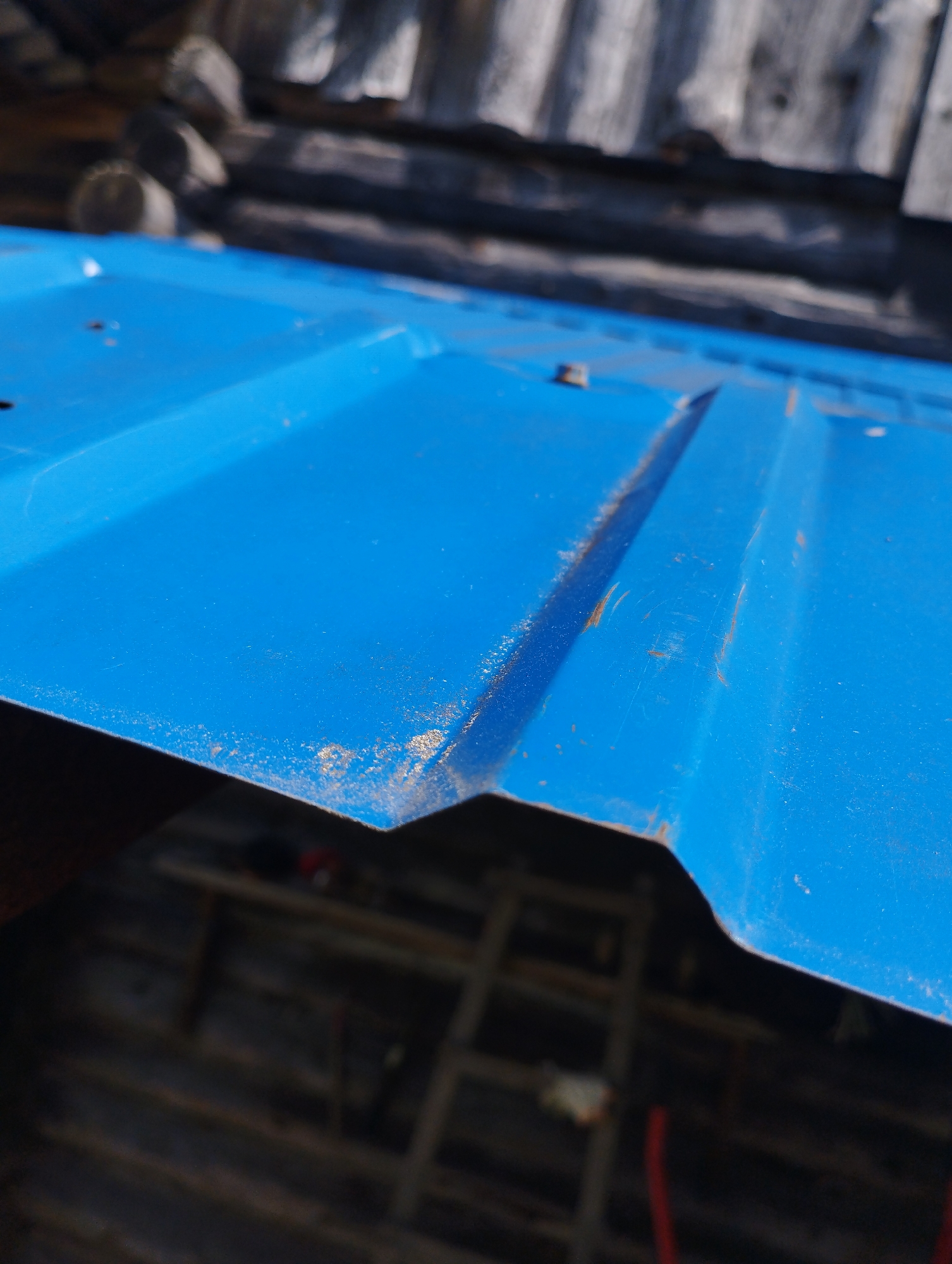 The image shows a close-up of a blue corrugated metal roof with some dirt and rust visible along the edges and seams. In the background, there is a blurred view of a wooden structure, possibly a roof or wall made of logs. The environment appears to be outdoors, under natural light. There are no people or specific actions depicted. The location is identified as Ширяево (ильинский), Russia.