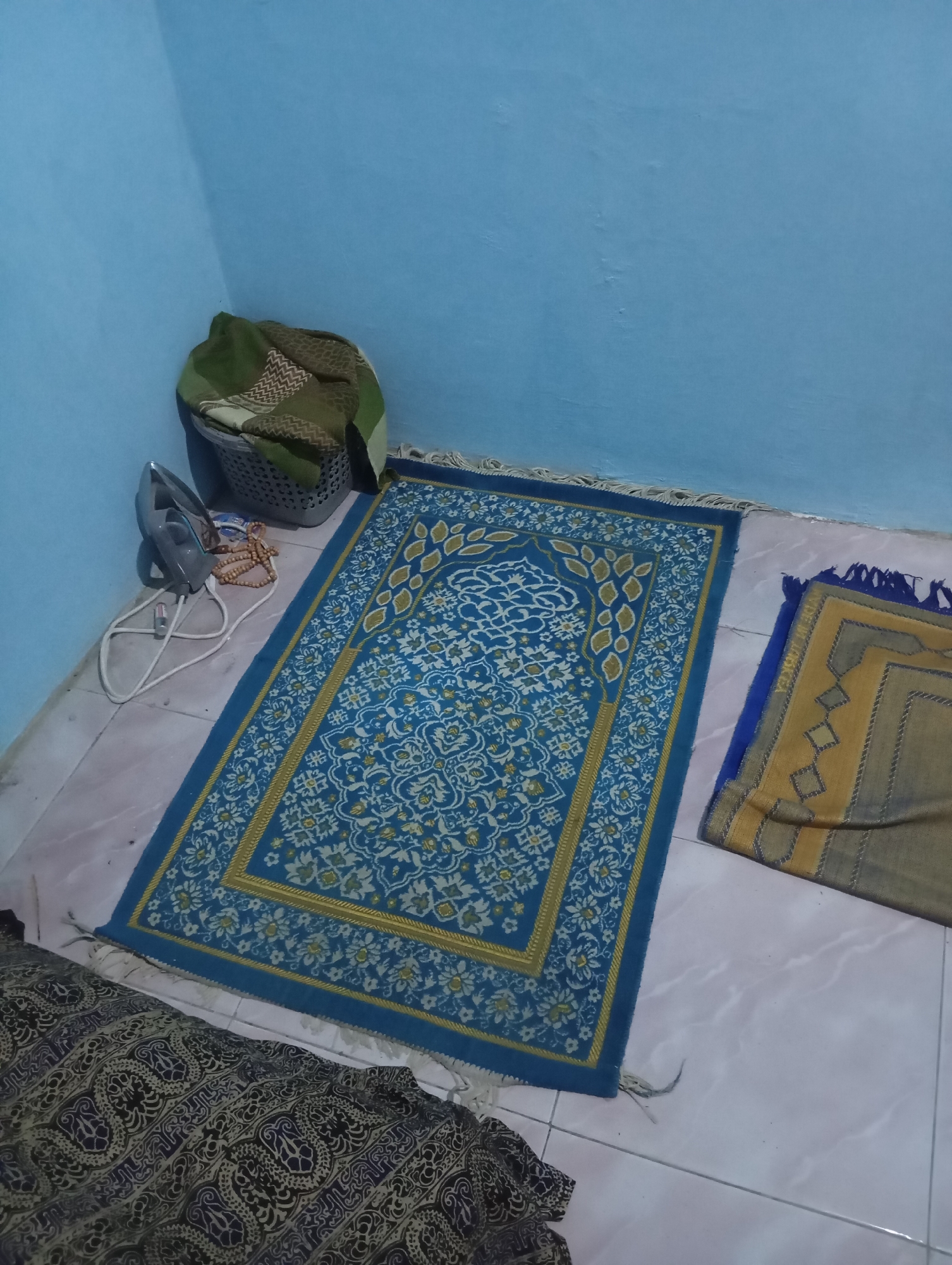 In a corner of a room with light blue walls, two prayer rugs are laid out on a tiled floor. The larger rug, predominantly blue with intricate white and yellow floral patterns and a gold border, is positioned centrally. To its right, a smaller, folded rug with a yellow and gray geometric design is visible. In the background, a grey plastic laundry basket contains folded clothing, and to the left of the basket, a silver electric iron rests on the floor beside a string of brown prayer beads. A dark fabric with a complex black and white pattern is partially visible in the foreground. The scene appears to be indoors, likely a domestic setting in Sudikampiran, Indonesia.