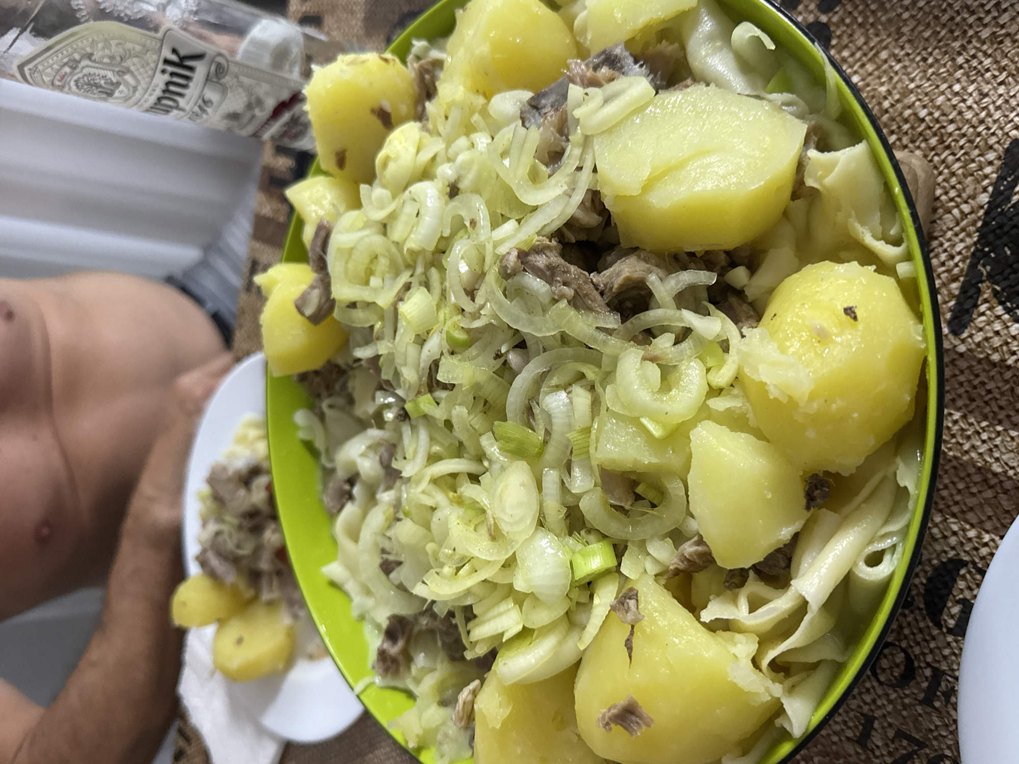 A close-up, slightly overhead shot shows a bowl of food, likely a meal, set on a patterned tablecloth. The bowl itself is a vibrant lime green and is overflowing with a hearty mix of ingredients. Dominating the composition are large, yellow, boiled potatoes, some cut in half, nestled amongst shredded white and green vegetables, possibly onions and leeks, and chunks of cooked meat. The food appears rustic and home-cooked. In the background, a person's bare torso is visible, suggesting they are seated at the table. To the right of the bowl, a clear glass bottle with a white and black label, partially filled with a clear liquid, stands upright. A white plate with a few boiled potatoes and meat is also visible in the upper left corner. The lighting is natural, and the overall impression is of a casual meal setting.