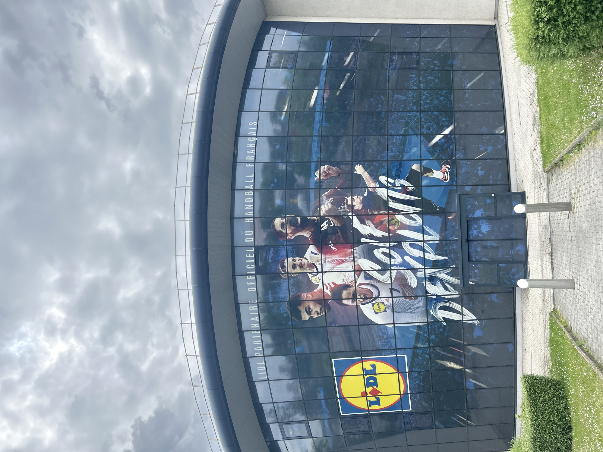 The image captures a building facade in Strasbourg, France. The building features a curved architectural design, with a large window display adorned with an advertisement. The advertisement showcases a collaboration between Lidl and the French Handball Federation ("LIDL PARTENAIRE OFFICIEL DU HANDBALL FRANÇAIS"), featuring images of handball players. Below the players is a clearly visible Lidl logo. The sky is overcast, and adjacent to the building is a paved walkway with manicured greenery.