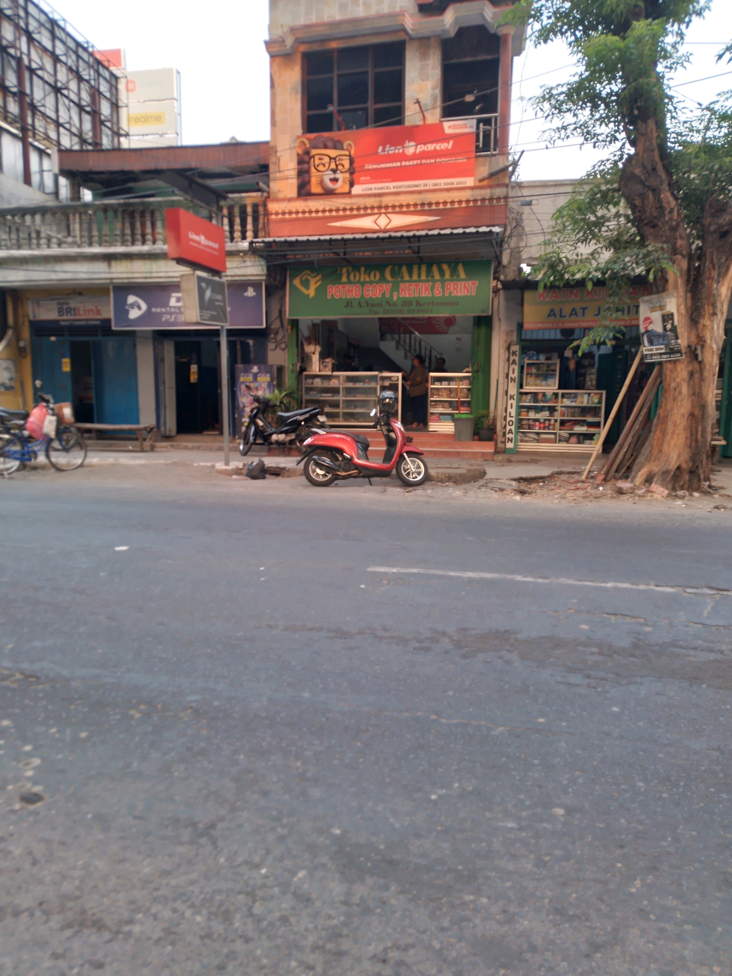 The image depicts a street scene in Kertosono, Indonesia, with several storefronts lining the right side of a paved road. On the left, a blue building with a BRI Link sign and blue shuttered windows is visible. Adjacent to it is a building with a red sign that reads "Lion Parcel," featuring an illustration of a lion wearing glasses. Next door, a green-awning storefront displays the text "Toko Cahaya / Foto Copy, Ketik & Print," with a telephone number provided below. Further to the right, a store with yellow signage announces "Kain Kilooan / Alat Jahit." Inside these shops, shelves stocked with various goods are visible. A red scooter is parked in front of the "Toko Cahaya" store, and a bicycle is parked near the BRI Link establishment. A large tree stands to the right of the shops, its trunk gnarled and weathered. The street itself is asphalted and appears to be moderately trafficked, with visible cracks and patches. Power lines crisscross above the buildings, and the overall lighting suggests it may be late afternoon or early morning.
