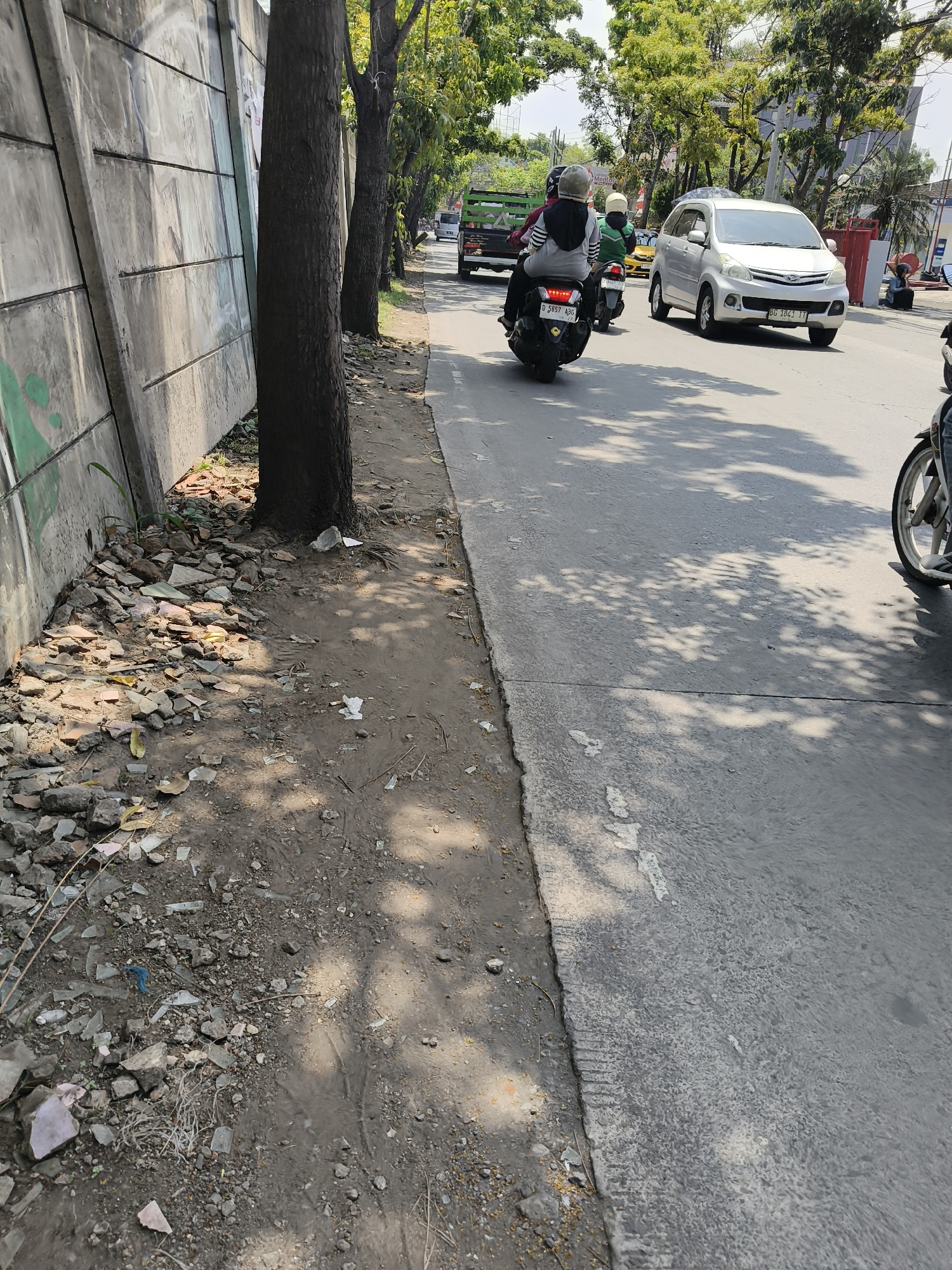 The image depicts a street scene in Bandung City, Indonesia, likely during the day under a clear sky, evidenced by the dappled sunlight and shadows on the road. The left side of the frame features a concrete wall, partially covered in graffiti, bordering a dirt path littered with debris, including broken pieces of tiles and glass. Several trees line the edge of the road, casting shadows on the pavement.
The road itself is a paved asphalt surface, marked with a white line. Traffic is present, with a silver minivan (license plate BC 1841 LT) visible in the mid-ground, driving in the same direction as the viewer. In front of the minivan, a motorcycle with two occupants wearing helmets is also in motion. Another motorcycle is partially visible in the lower right corner of the image. Further in the distance, a truck with a green cargo bed is also present on the road. The presence of multiple vehicles suggests this is a moderately trafficked urban road. The overall environment appears to be a busy street with a mix of infrastructure and natural elements.