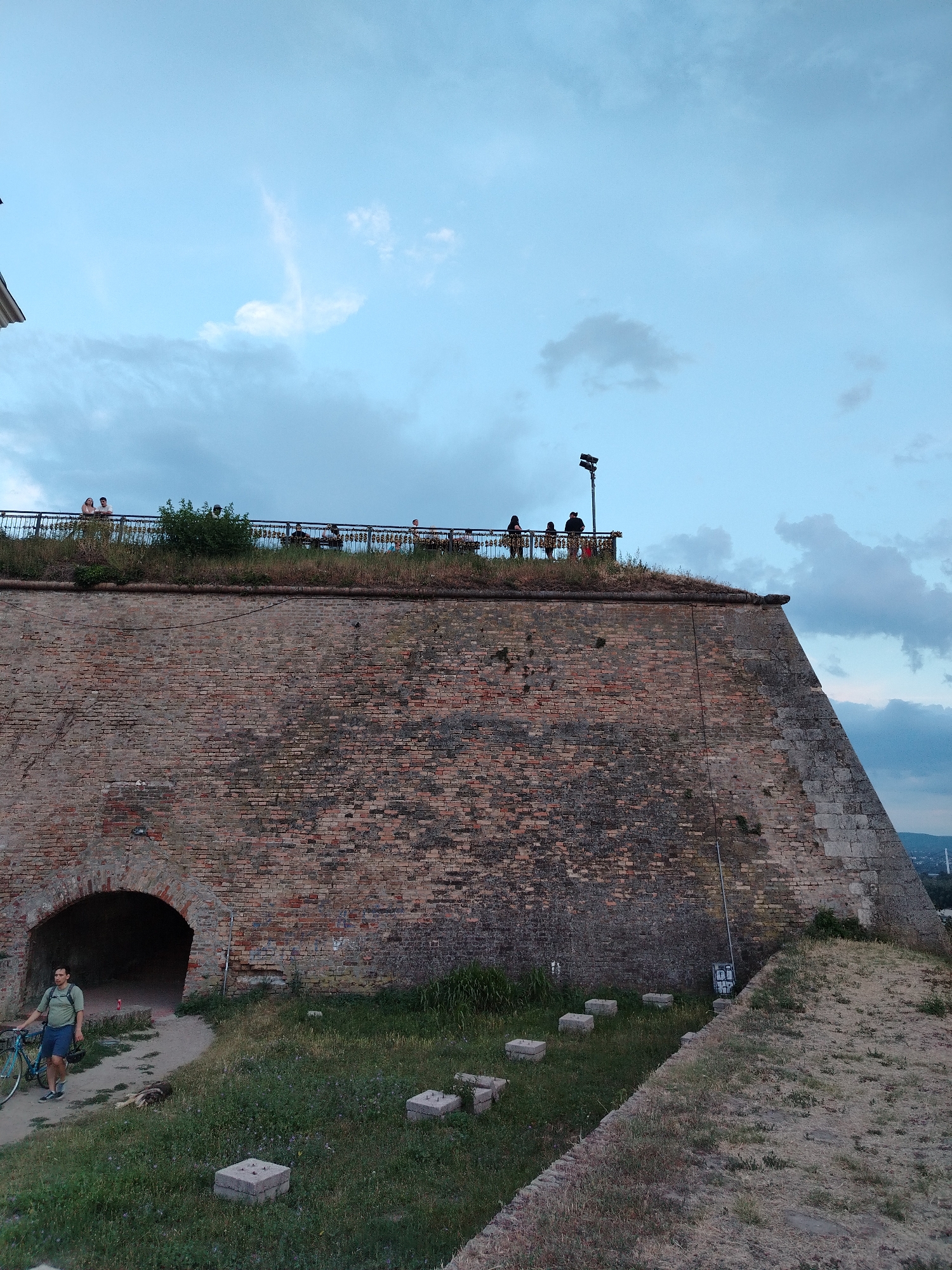 In Petrovaradin, Serbia, a wide shot captures a large, ancient brick fortress wall with an arched opening. The wall dominates the foreground and mid-ground. On top of the wall, a railing separates a grassy area where several people are standing and observing the surroundings. One person, a male wearing a green shirt and blue shorts, walks with a bicycle near the arched opening. The sky above is a pale blue with scattered clouds, indicating it is likely late afternoon or early evening. The environment appears to be an outdoor historical site.