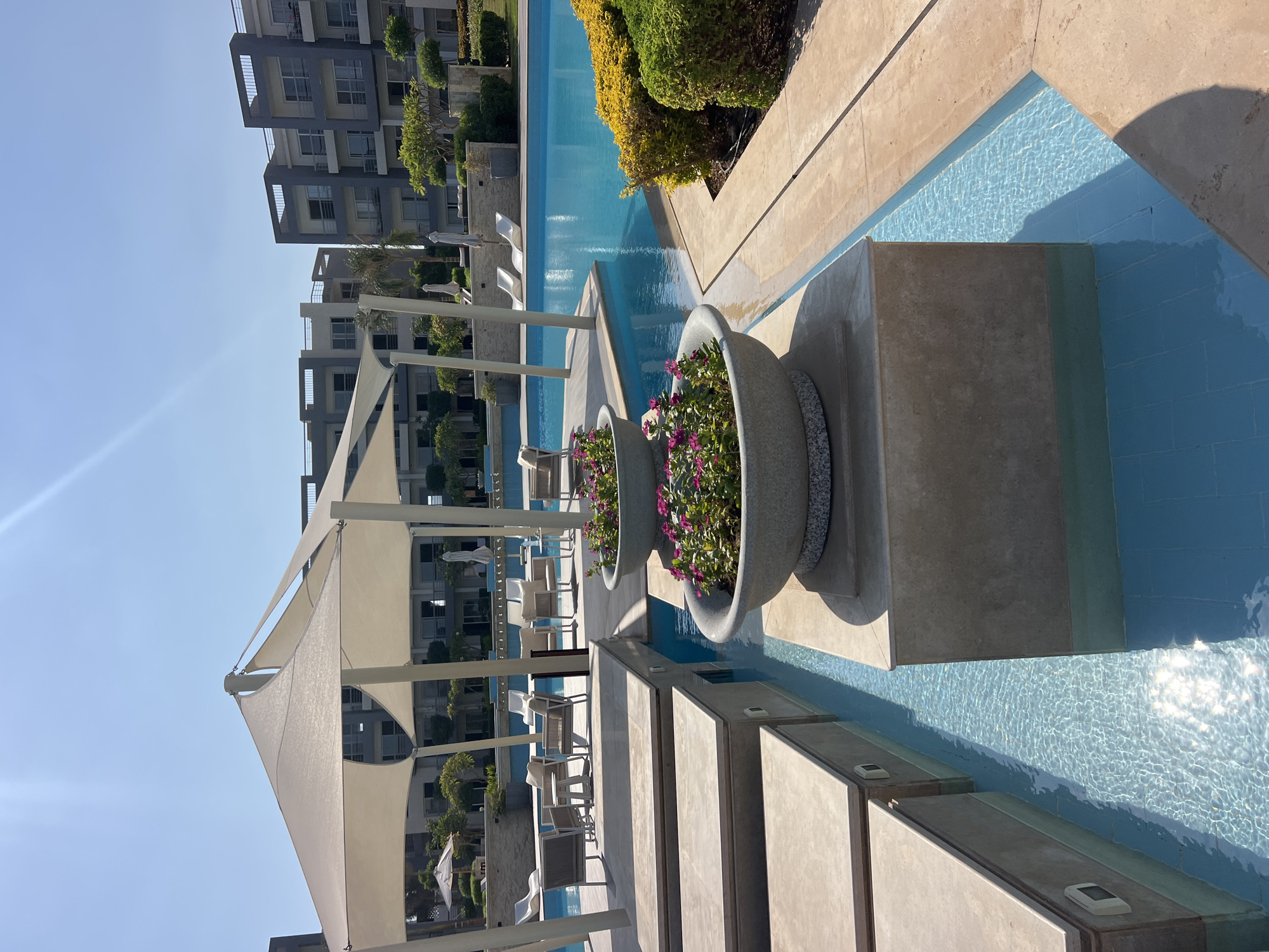 The image depicts a resort scene with a swimming pool and modern buildings in the background. In the foreground, there are two large planters filled with pink flowers, sitting on a raised stone structure. A shallow water feature with blue tiles runs alongside the planters, reflecting the sunlight. Beyond this, a large, turquoise swimming pool is visible, surrounded by lounge chairs and umbrellas. The architecture of the buildings is contemporary, with multiple stories and balconies. Lush green landscaping, including bushes and trees, complements the scene. The sky is clear and blue. The overall impression is one of a luxurious and relaxing resort environment. The location is identified as Unknown, Egypt.