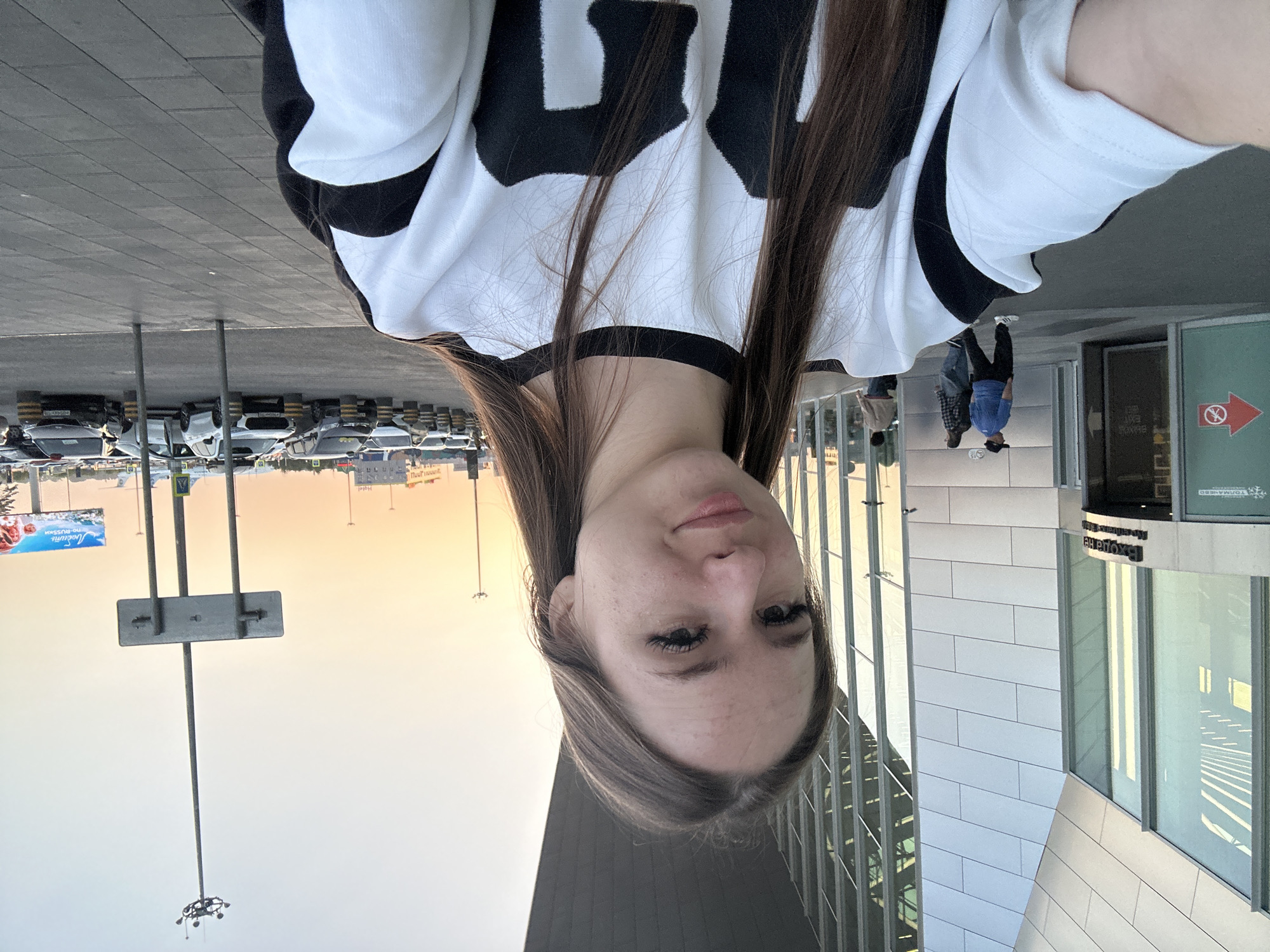 A young woman with long brown hair is taking a selfie outdoors in front of a modern building. She is wearing a black and white sweatshirt. Behind her, there are cars parked in a lot, and in the distance, an airplane can be seen. The sky is a pale orange, suggesting it is either sunrise or sunset. The building has large glass windows and a metallic facade. There are a few other people visible in the background walking near the entrance of the building. A large billboard is visible in the far right distance.