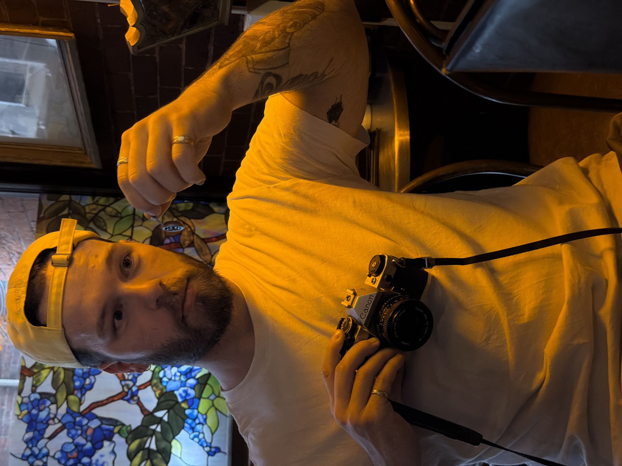 A man stands in a dimly lit interior space with a stained-glass window visible in the background, possibly in Boston, Massachusetts, USA. He wears a light-colored t-shirt and a yellow baseball cap. He has a short beard and is holding a vintage Canon film camera. He has tattoos on his right arm and is wearing a ring on his finger in each hand. Part of a dark-colored chair is visible in the background, along with what appears to be brickwork and a framed picture. The lighting casts a warm, yellow hue over the scene.