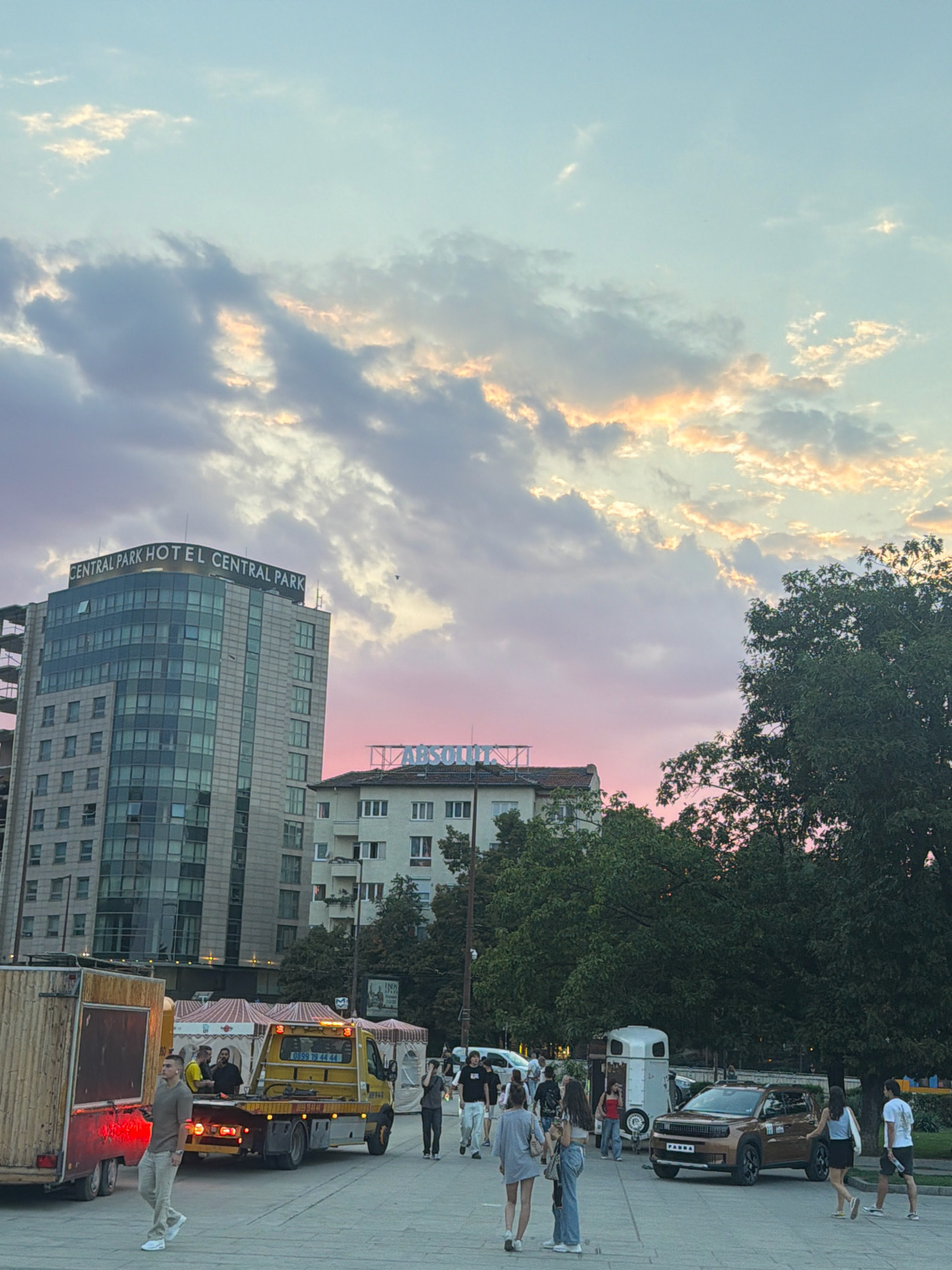 In the city of Sofia, Bulgaria, a street scene unfolds under a sky painted with soft pink and orange hues, indicative of dusk. Dominating the skyline is the "Central Park Hotel," a modern, multi-story building with a glass facade. Adjacent to it, a lower, older building displays a prominent sign with the word "ABSOLUT". In the foreground, a street scene is depicted with various elements. A yellow tow truck, bearing the phone number "0899 79 44 44", is positioned in the middle ground, with its platform extended. To its left, a large, wooden trailer with red lighting is parked. Several people are visible walking on the paved area, some appearing to be interacting with each other. A brown SUV with a license plate that reads "PARKA" is parked to the right, near a small white trailer, possibly for horses. Lush green trees provide a natural backdrop to the urban setting. The overall impression is a dynamic urban environment during a transitional time of day, with a mix of modern and older architecture.