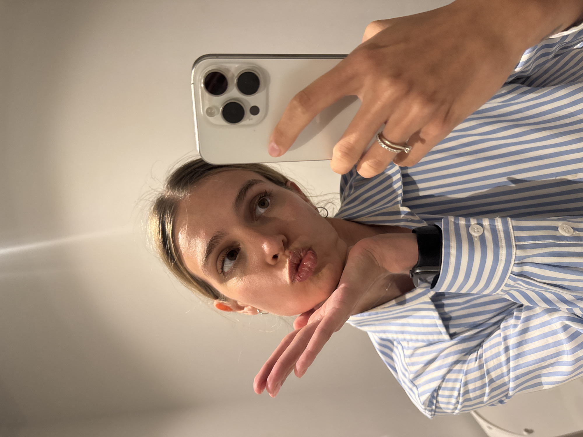 A woman is taking a mirror selfie, with the image presented at a 90-degree clockwise rotation. She occupies the lower-right portion of the frame, with her head tilted as she looks upwards and to the right. Her light brown hair is visible, and she is making a "kiss" face with pursed lips. Her left hand is raised, with fingers gently curled, resting playfully under her chin.
Her right hand holds a white or silver iPhone, which is prominently displayed across the top-center of the frame. The smartphone is encased in a clear cover, and its triple camera system is clearly visible on the back. She is wearing a blue and white striped collared shirt. A silver ring with multiple bands is on her right ring finger, and a black smartwatch is visible on her left wrist. The background consists of a plain, light-colored wall, indicating an indoor setting with soft, ambient lighting. No text is visible in the image. The overall impression is casual and playful.
