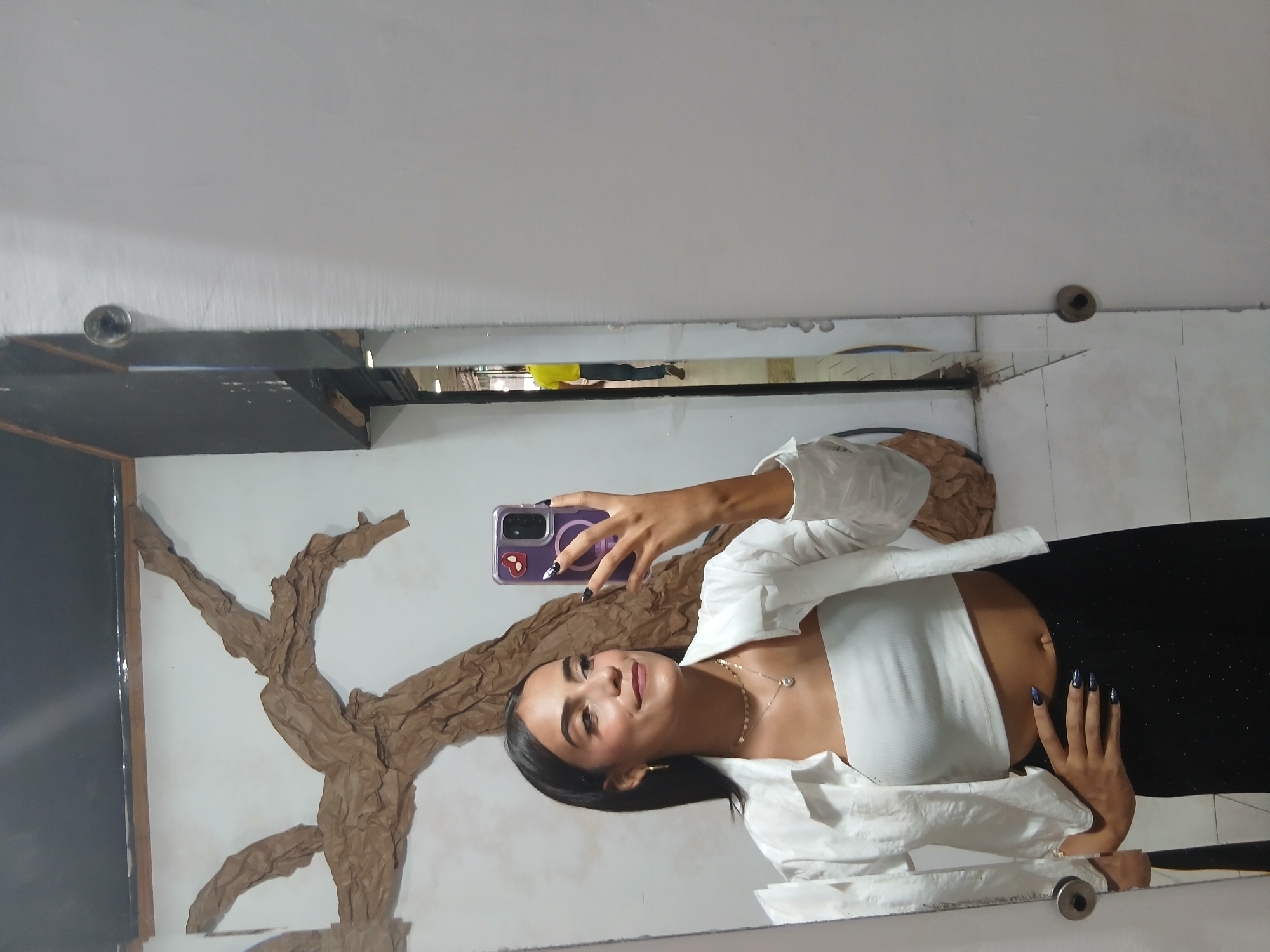 A young woman is taking a selfie in a mirror. She is wearing a white top under a white open-front shirt and black pants. Her hair is dark, and she is wearing makeup and jewelry. She is holding a purple smartphone in her right hand. In the background is a white wall with a brown tree-like decoration. The setting appears to be indoors, possibly in a home or apartment. The location in Porlamar, Venezuela, is not evident from the image.