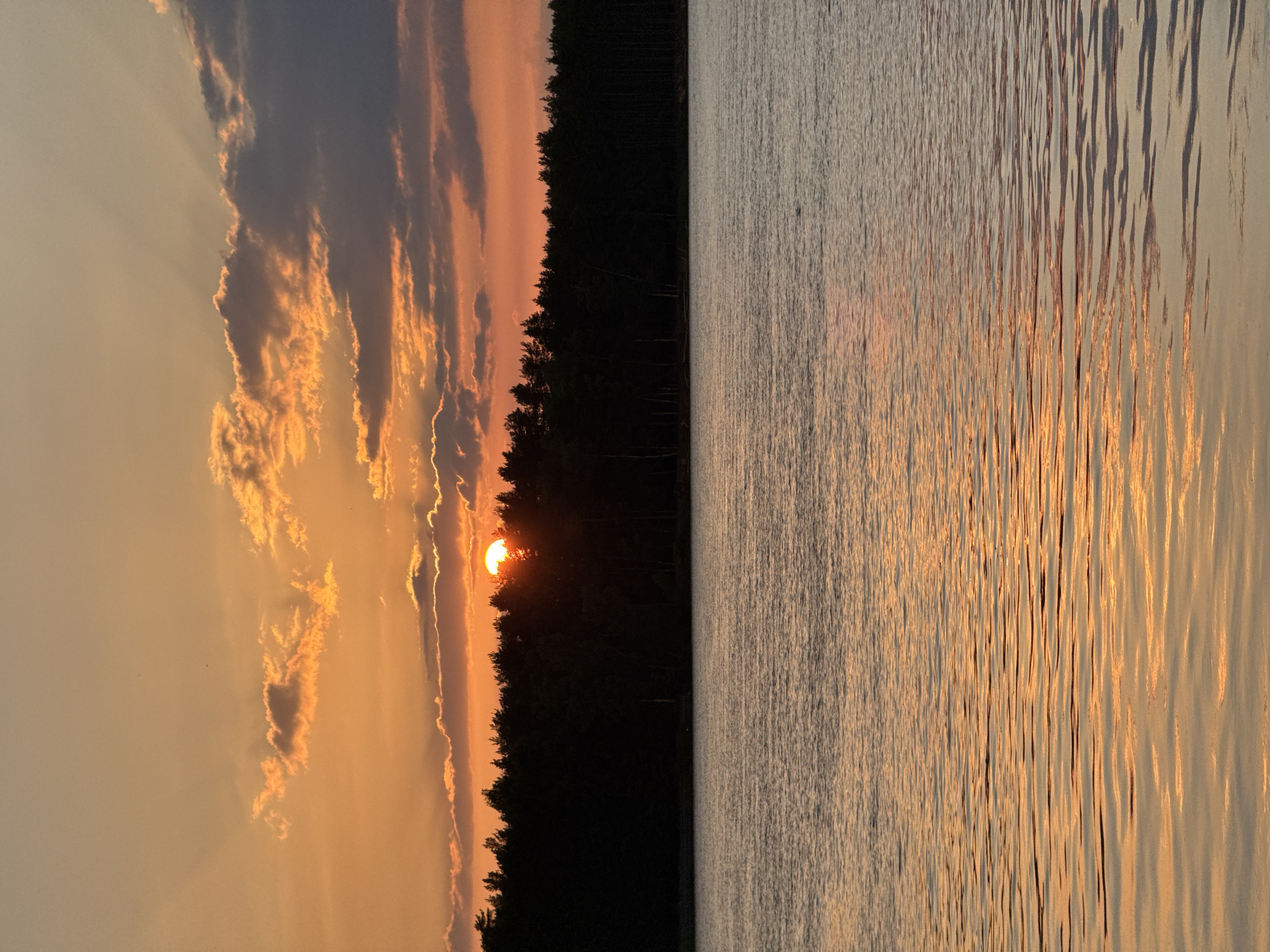 The image depicts a sunset over a body of water, with a dense forest lining the far shore. The sun, partially obscured by clouds, casts a warm orange and yellow glow across the sky and reflects on the rippling surface of the water. The sky is filled with dramatic clouds, some dark and heavy, others tinged with the sunset's light. The silhouette of the trees creates a stark contrast against the vibrant sky. The scene appears to be a natural landscape, likely a lake or a wide river. The image captures a tranquil and picturesque moment in nature, characteristic of the landscapes found in and around Carnikava, Latvia, a region known for its coastal and riverine scenery. No people or specific man-made structures are clearly visible in this particular framing of the scene.