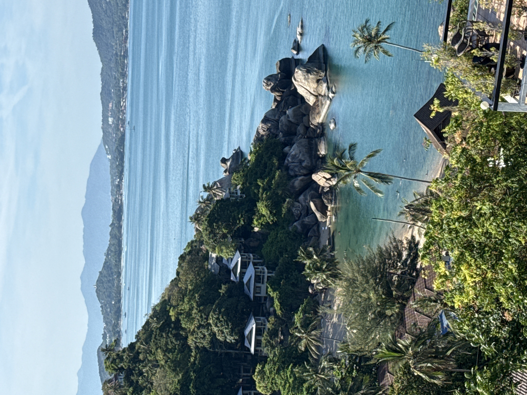 The image presents an aerial view of a tropical landscape, potentially located in Baan Chaweng Noi, Thailand, given the context. The scene is dominated by a rocky promontory jutting into a turquoise sea. Large granite boulders cluster at the tip of the promontory, surrounded by dense green vegetation including various trees and palms. Several buildings with white walls and distinct architectural features are nestled within the lush foliage. The sea stretches out towards a distant coastline, which appears as a hazy blue outline against the sky. The overall impression is one of a tropical paradise.