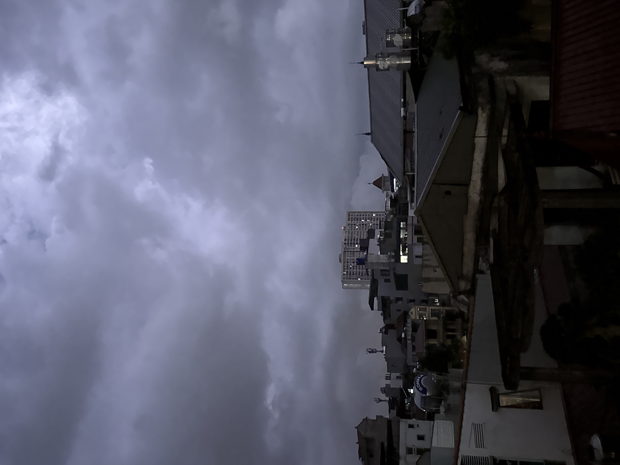 The image shows an overcast cityscape, likely in Hanoi, Vietnam. A tall, modern building rises in the center-left distance amidst older, low-rise structures. In the right foreground, the dark rooftops of adjacent buildings are visible. The sky is predominantly gray and filled with heavy clouds.