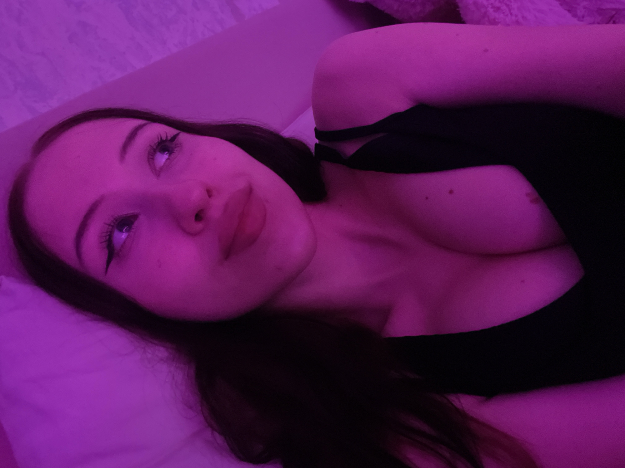 The image shows a close-up, indoor shot of a young woman with dark hair, wearing a black top. She is illuminated by pinkish-purple light. Her head is tilted, and she is looking off to the side with her eyes open. She has on dramatic winged eyeliner and full lips. The background is blurred, but appears to be a bed with a textured headboard. There is no indication of the city of Votkinsk, Russia in the image.