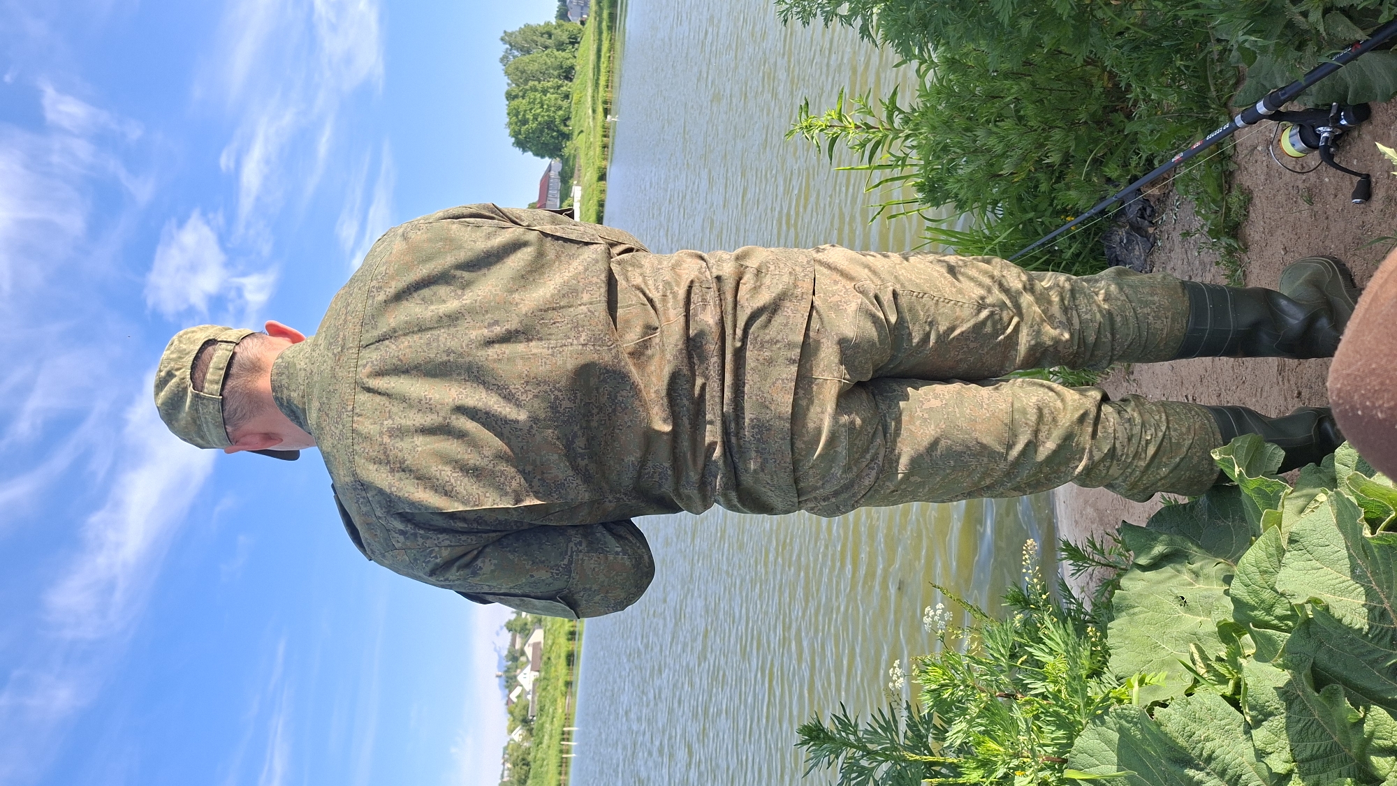 A man, seen from behind, stands on the bank of a body of water under a bright blue sky dotted with wispy white clouds. The image is captured on a clear, sunny day. He is dressed in full camouflage gear, comprising a long-sleeved shirt, trousers, and a cap, all featuring a pixelated green and brown pattern. He is also wearing dark green rubber boots. The man appears to be looking down, possibly at the water or his equipment.
To his right, a fishing rod with a reel and bright yellow fishing line rests on the ground beside the water, indicating a fishing activity. Text visible on the fishing rod includes "GO FEEDER" and "CALIBER GO". The immediate bank features green vegetation, including large-leaved plants in the foreground, and exposed dirt.
The body of water, likely a lake or wide river, extends into the distance. On the far bank, a treeline and several distant buildings, possibly houses, are visible, suggesting a rural or natural setting. The overall scene depicts a calm outdoor pursuit. The location context for this image is Забалоцкі сельскі Савет, Belarus.