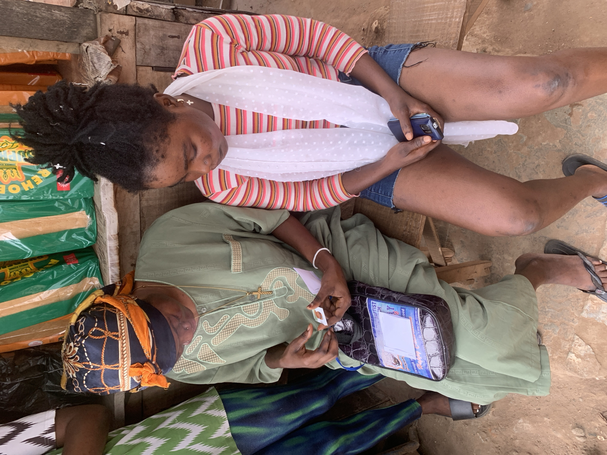 The image depicts two women sitting on a wooden bench in an outdoor market setting, likely in Shagamu, Nigeria. The woman on the left wears a green garment and an orange and black headscarf. She is holding a small, light-colored card in her hands, examining it. A dark, textured bag with a clear plastic pouch is placed in front of her. The woman on the right has dark, braided hair and wears a striped shirt and denim shorts. She is looking down and holding a dark-colored mobile phone. A white, patterned scarf is draped around her neck. The ground is dirt, and in the background, there are bags of goods, some with green and orange packaging. The overall scene suggests a moment of transaction or information exchange in a local marketplace.