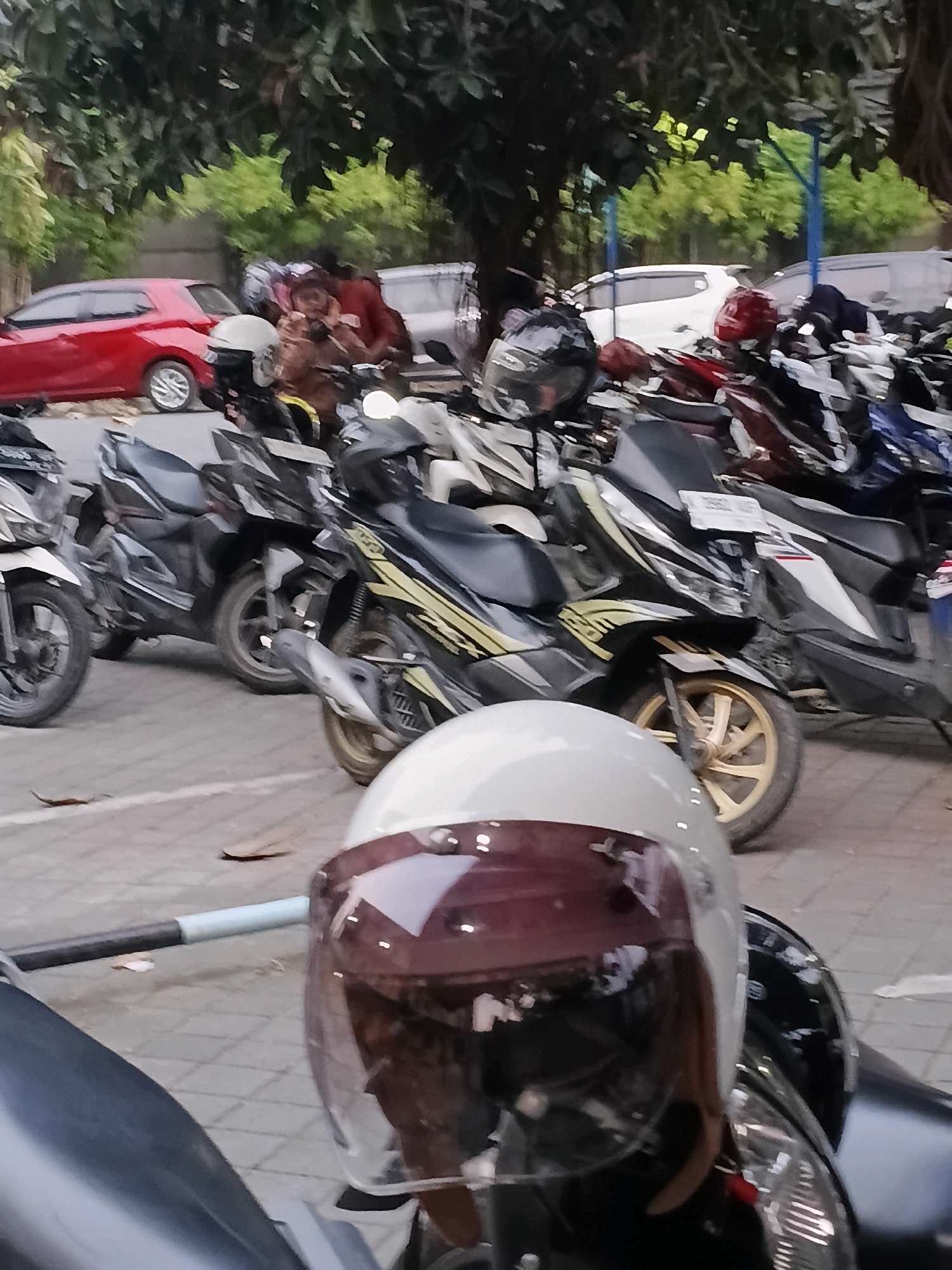 In the city of Purwodadi, Indonesia, a parking lot is depicted filled with numerous parked motorcycles and a few cars. The foreground prominently features the front of a white motorcycle with a burgundy and transparent visor. Behind it, several other motorcycles are visible, some with black fairings and yellow accents, and others in various colors like red and blue. Two people are seated on a motorcycle in the midground, one wearing a brown jacket and the other a darker outfit. A red car is parked on the left side of the image, with a white car and other vehicles further in the background. Trees with green foliage line the upper portion of the scene, suggesting an outdoor setting with some shade. The ground is paved with interlocking concrete blocks.