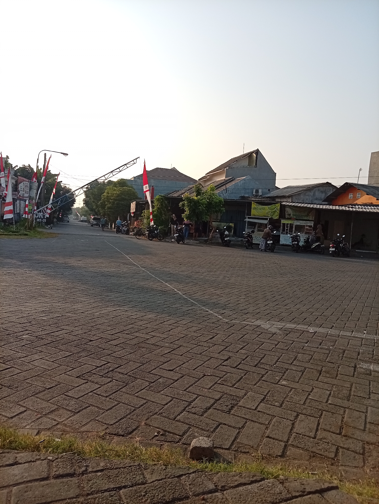 This image depicts a street scene in Bligo, Indonesia, possibly near the intersection of Jalan Bligo and Jalan Raya Gondangrejo. The foreground is dominated by a paved street with interlocking bricks arranged in a herringbone pattern. A strip of dry grass and a few small stones line the edge of the pavement.
On the left side of the street, a lamppost stands tall, adorned with several Indonesian flags (red and white stripes). A large, angled metal structure, possibly a temporary stage or support for decorations, extends across the street. Further down the street on the left, there are buildings with signage, indicating commercial activity, and several parked motorcycles. A few people are visible walking and standing near these buildings.
The right side of the street features a row of shops and food stalls, also with some Indonesian flags displayed. More parked motorcycles are lined up in front of these establishments. Several individuals are interacting near the stalls, including one person appearing to be purchasing food. The background shows residential buildings and trees under a bright, clear sky, suggesting it is daytime, likely morning or late afternoon due to the soft lighting. The overall atmosphere is that of a quiet, developing urban or suburban area.