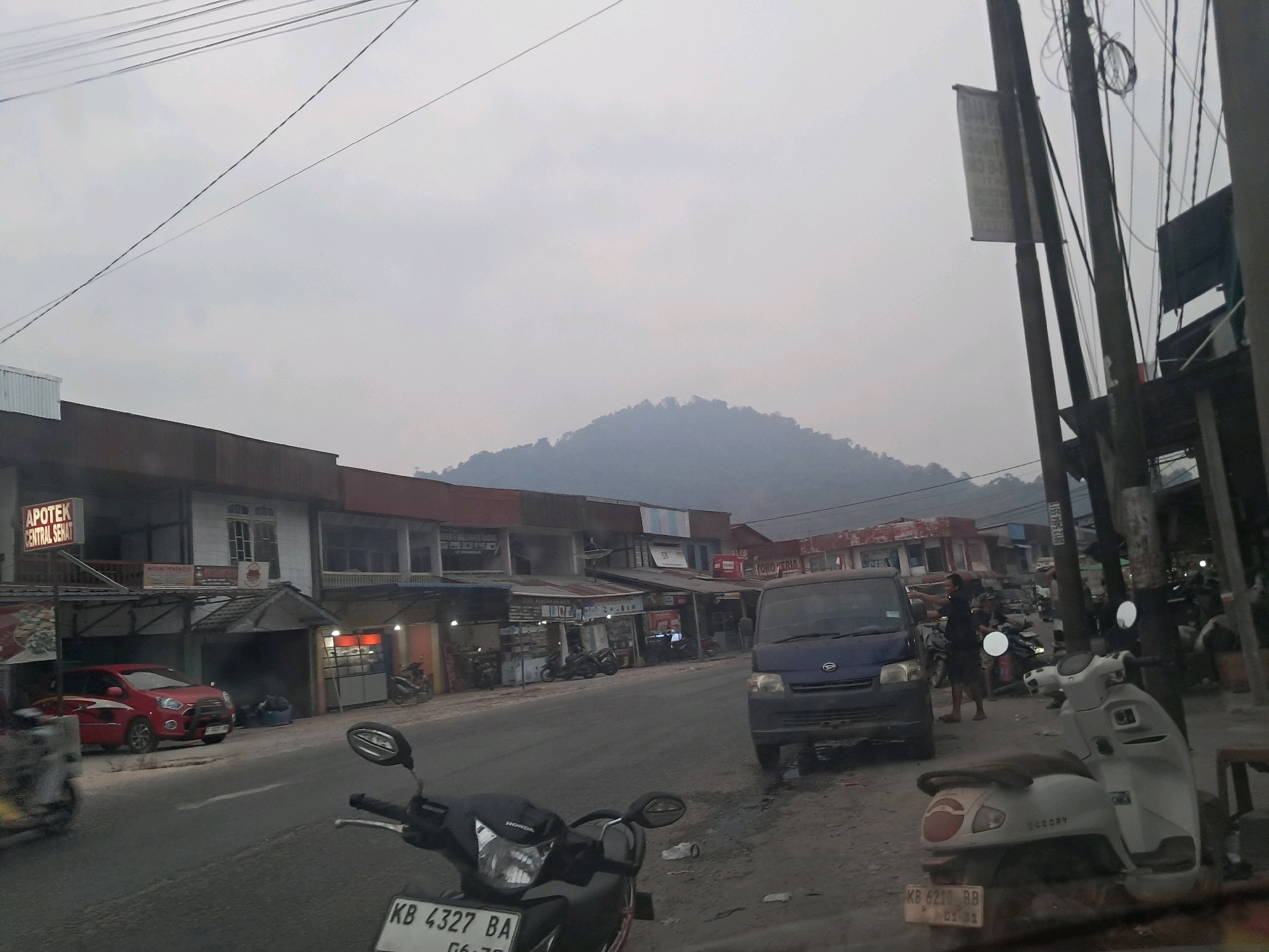 This image captures a street scene in Anjungan Melancar, Indonesia, likely during overcast conditions or early morning/late evening. The road is paved, with a shoulder of dirt and gravel. On the left side of the road, a row of low-rise buildings with storefronts lines the street. One building prominently displays "APOTEK CENTRAL SEHAT" on a sign. A red car is parked in front of one of these establishments. Further down the street, a dark blue van is parked, facing the viewer.
Several motorcycles are visible, some parked and others seemingly in motion. In the foreground, the handlebars and front wheel of a black motorcycle are prominent, with a license plate reading "KB 4327 BA". To the right, a white scooter with a visible license plate "KB 6210 BB" is parked. A person in a dark shirt is standing near the blue van, gesturing with their hands. Another person is seated on a motorcycle in the background. The sky is a muted grey, and a hazy, tree-covered hill or mountain range is visible in the distance, partially obscured by the haze. Electrical wires crisscross the sky above the buildings.