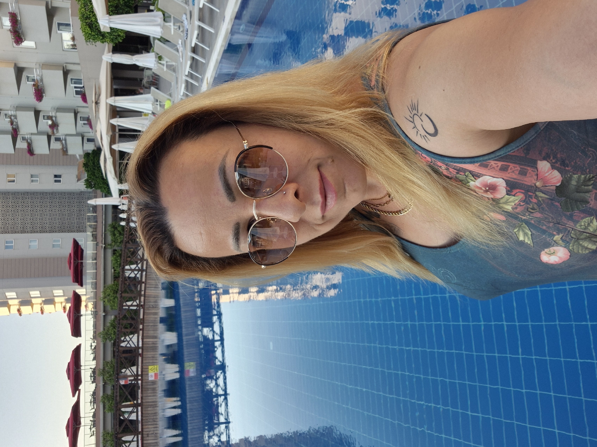 A woman with blonde hair and sunglasses is taking a selfie in a swimming pool. She is wearing a blue top with a floral pattern. A tattoo of a sun symbol is visible on her right shoulder. The background shows a hotel with balconies and red umbrellas by the pool. The water in the pool is blue and reflects the surrounding structures. The image appears to be taken during the daytime, with bright lighting.